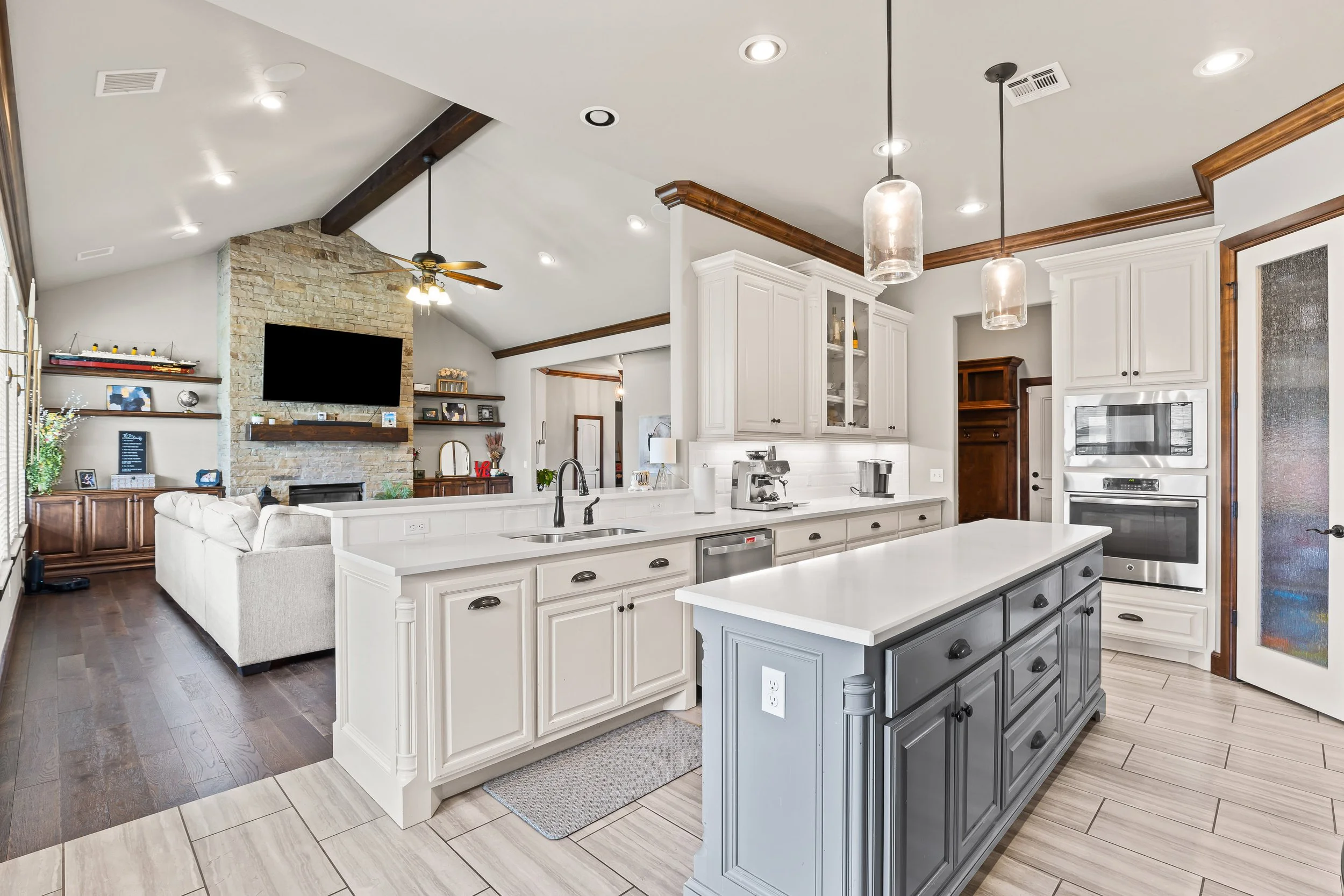 Open-concept kitchen with white cabinets, gray island, pendant lights, and living room with TV and shelves in background. Real estate photography in Yukon, OK.