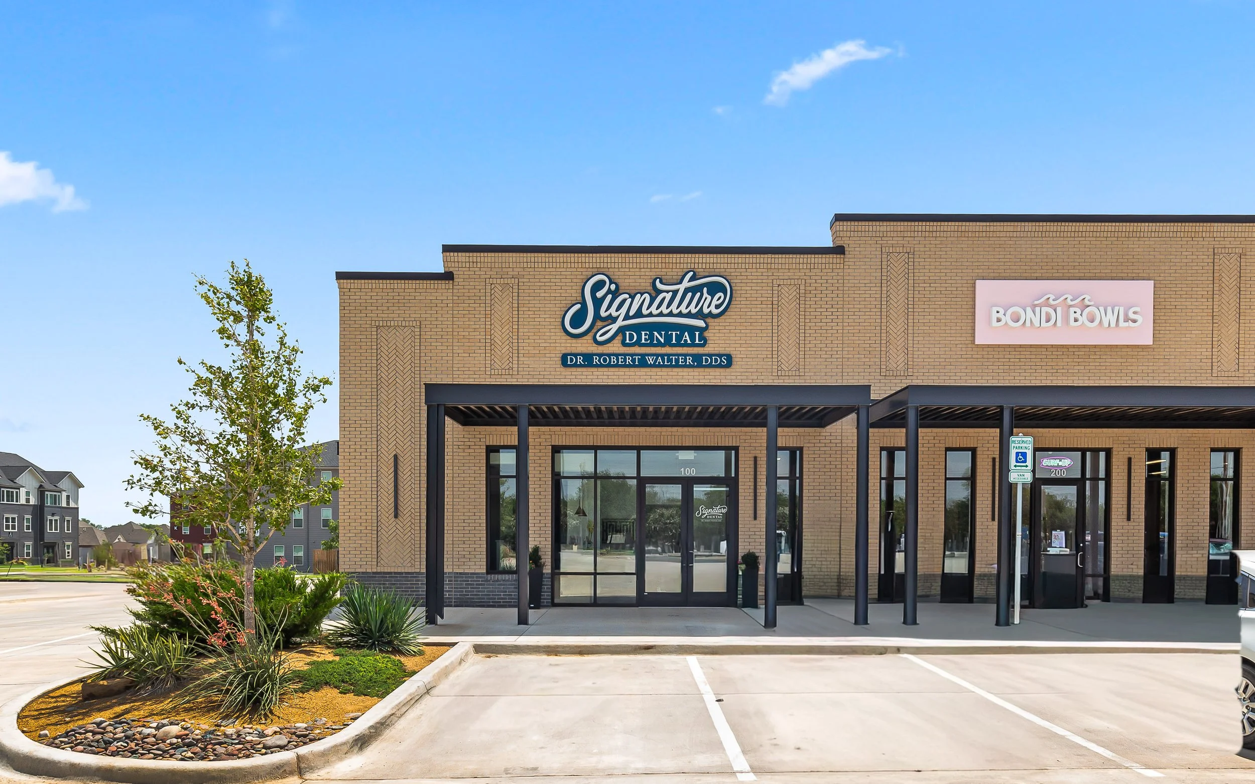 An empty parking lot in front of a brick building with signs for Signature Dental and Bondi Bowls, under a blue sky with some clouds.
