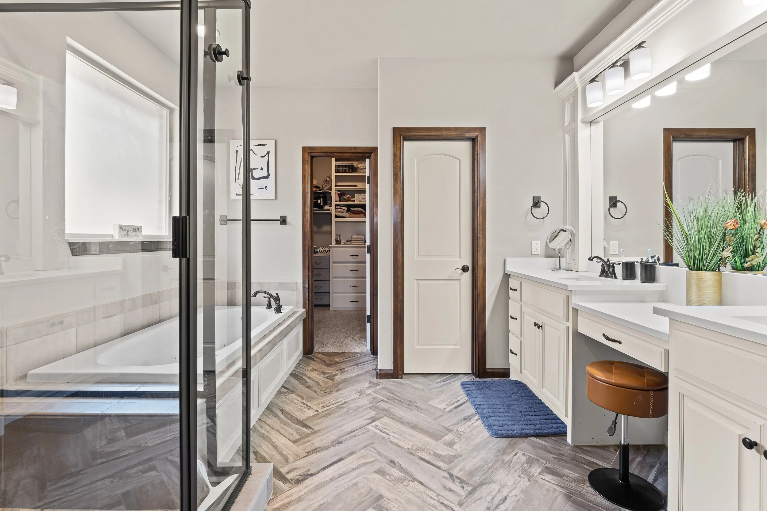 A spacious bathroom with a walk-in shower, bathtub, double sink vanity with a large mirror, and decor including a tall green plant and a round brown stool. Real estate photography in Yukon, OK.