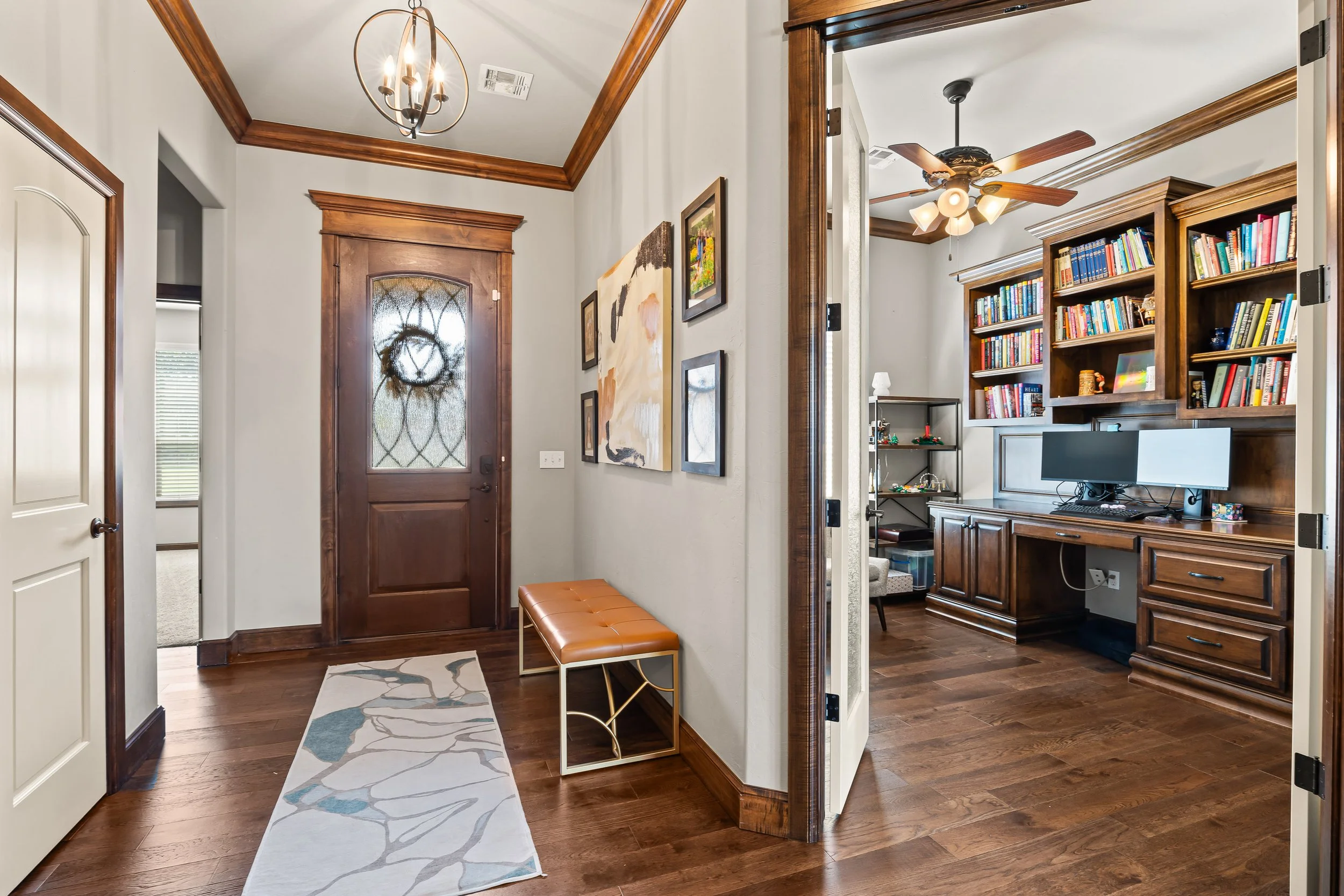 Entryway area with a wooden front door and a small tan bench with a gold metal frame. The hallway has white walls with art pictures and dark brown wooden trim. There is a light fixture with exposed bulbs hanging from the ceiling. To the right, a door