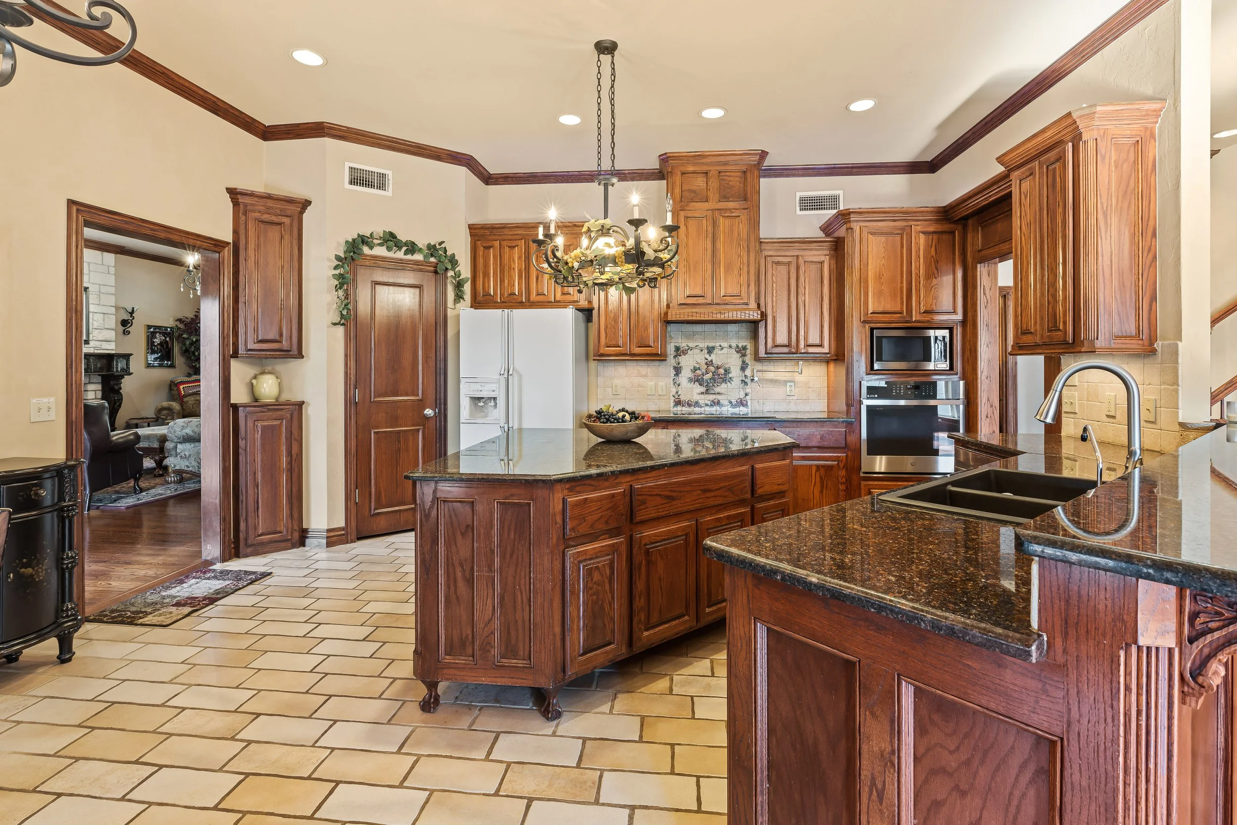 A kitchen with wooden cabinets, a kitchen island, a chandelier, and beige tiled flooring. Real estate photography in Mustang, OK.