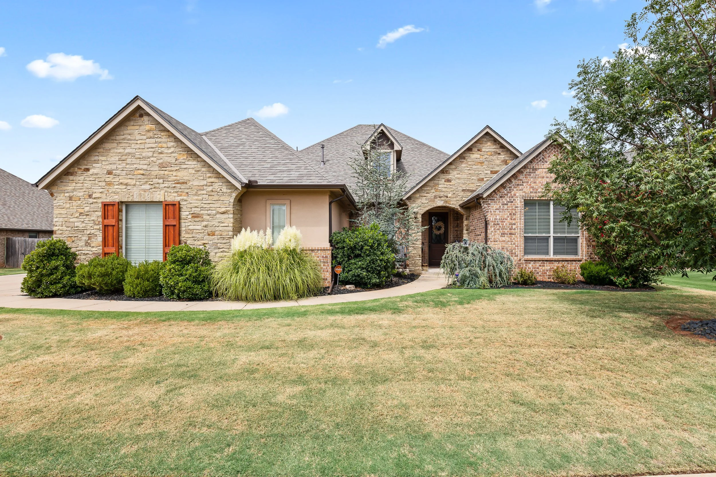 Front view of a suburban house with a brick and stone exterior, surrounded by a well-maintained lawn and various bushes, under a clear blue sky. Real estate photography in Yukon, OK.