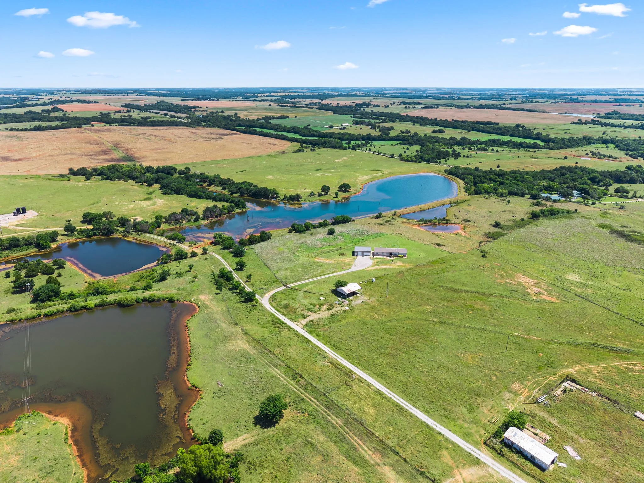 Aerial view of a rural landscape with green fields, small lakes, scattered houses, and a road, under a blue sky with a few clouds. Drone photography in Ardmore, OK.