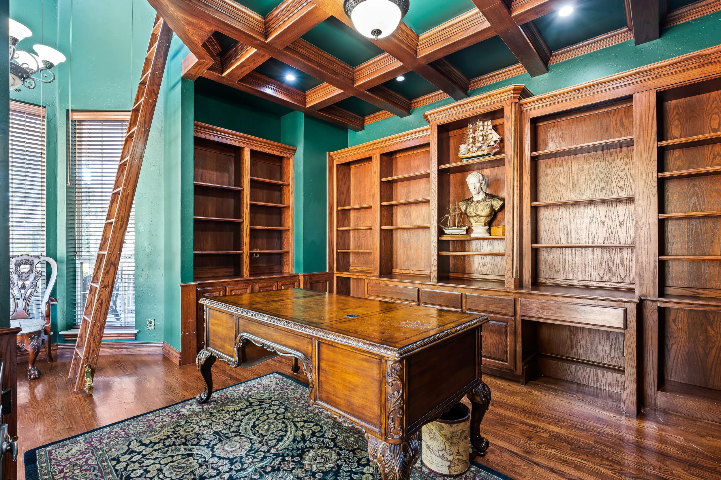 A wood-paneled office with built-in bookshelves, a wooden desk, a patterned area rug, and a wooden ladder leaning against the bookshelves. The room has a green ceiling with wooden beams and a window with blinds. Real estate photography in Mustan, OK.