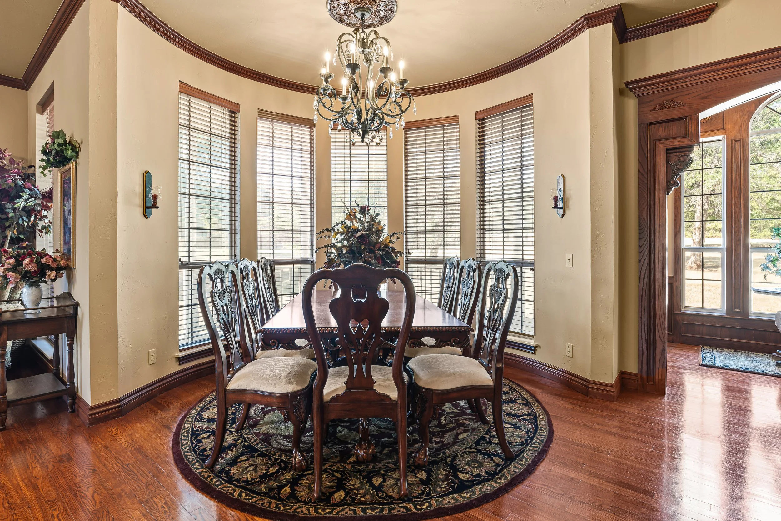 A dining room with a round table and eight wooden chairs, a chandelier hanging from the ceiling, large windows with blinds, and wooden trim and moldings, with a floral arrangement as a centerpiece. Real estate photography in Mustang, OK.