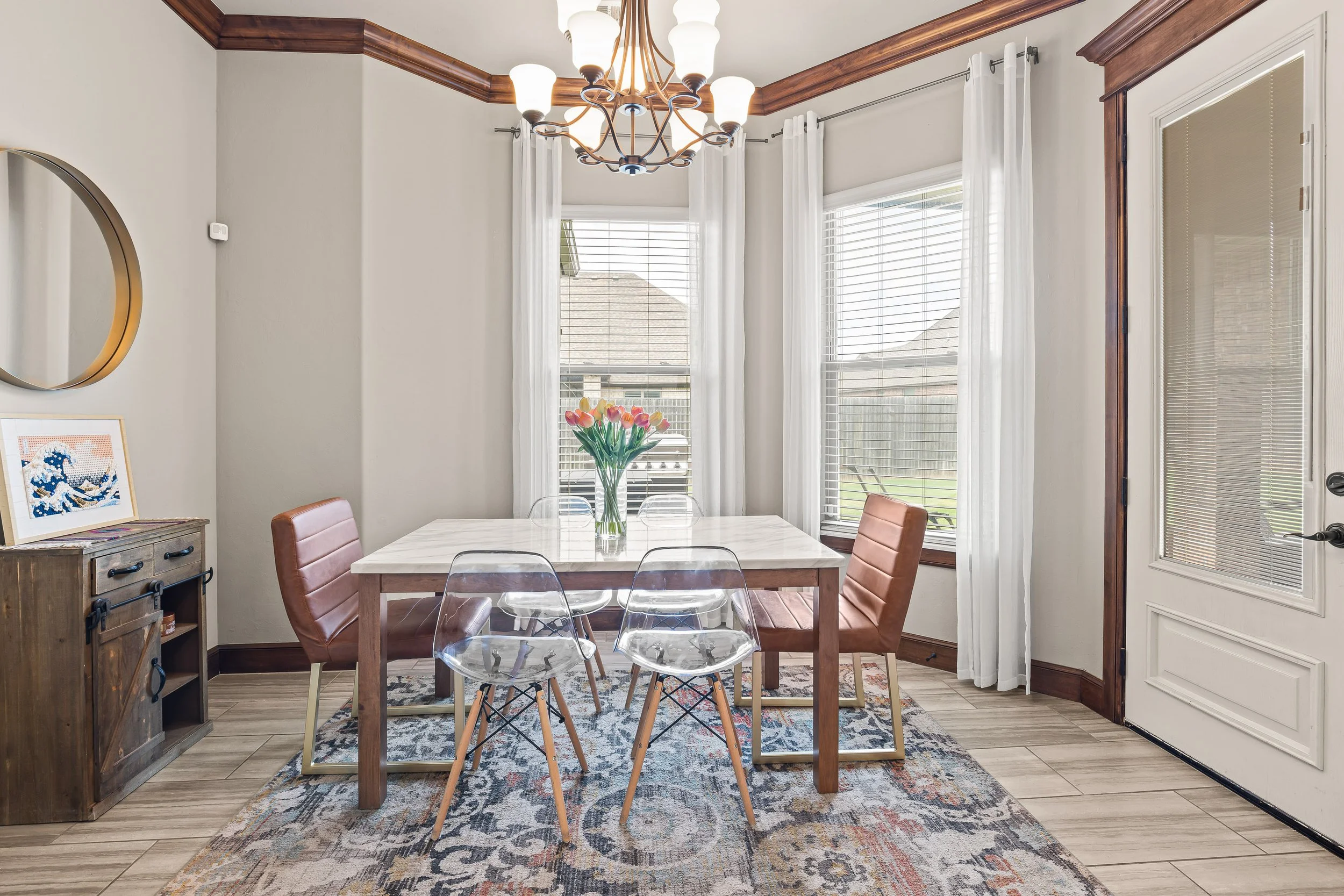 Dining room with a wooden table, pink upholstered chairs, and clear acrylic chairs, a vase of pink tulips, large windows with white curtains, a chandelier, a colorful area rug, and a small wooden cabinet. Real estate photography in Yukon, OK.
