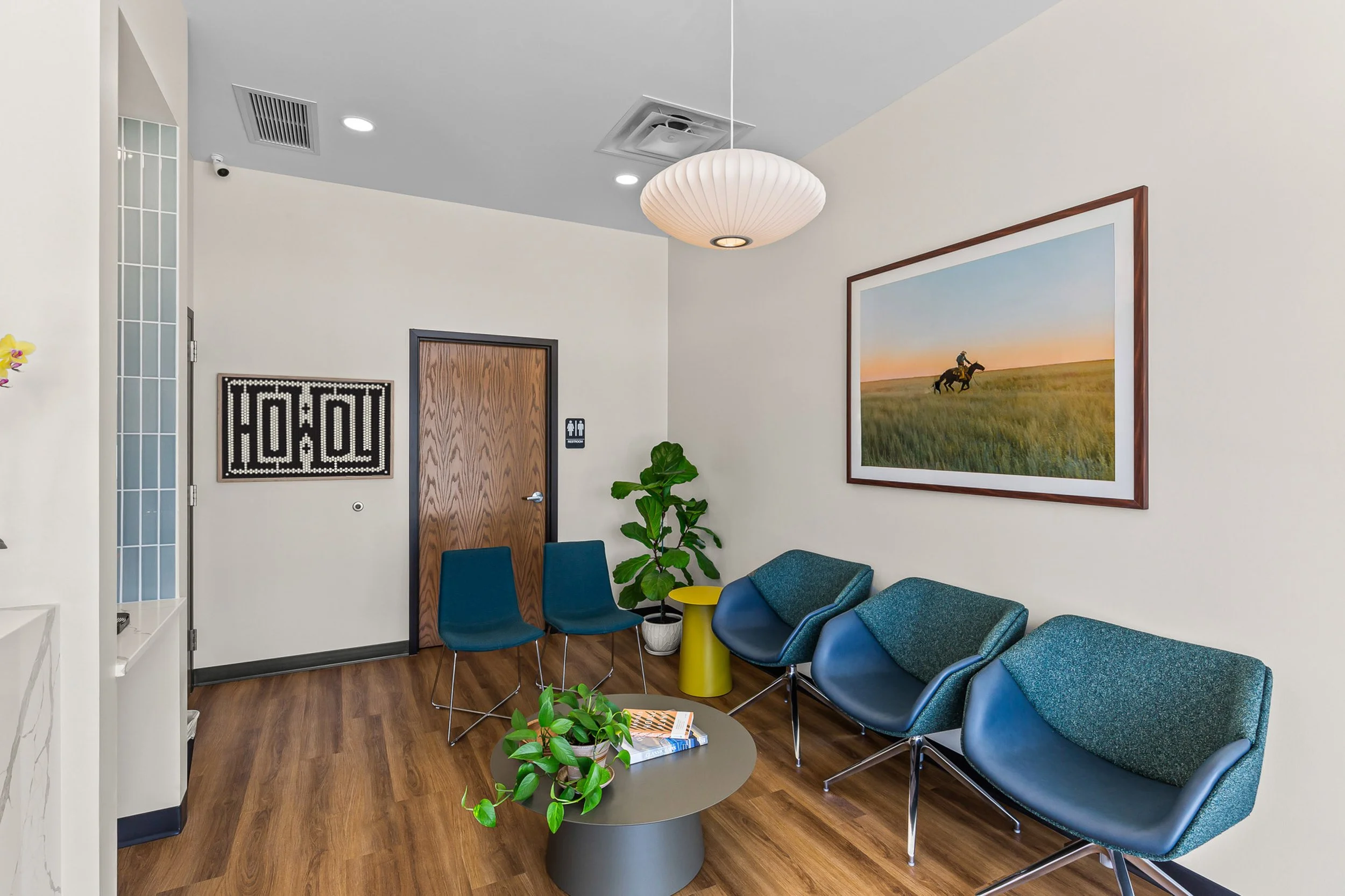 Waiting area with six blue chairs, a small yellow side table, potted plants, a large framed landscape photograph, and a round coffee table with magazines. Commercial Photography in Mustang OK.