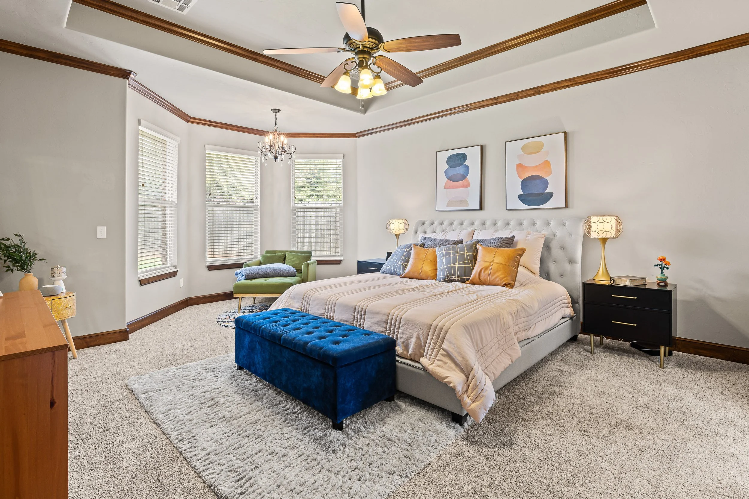 A bedroom with a large bed with beige bedding and multiple pillows, a blue velvet bench at the foot of the bed, two nightstands with lamps, three framed abstract art pieces on the wall above the bed, a green armchair with a blanket near three large w