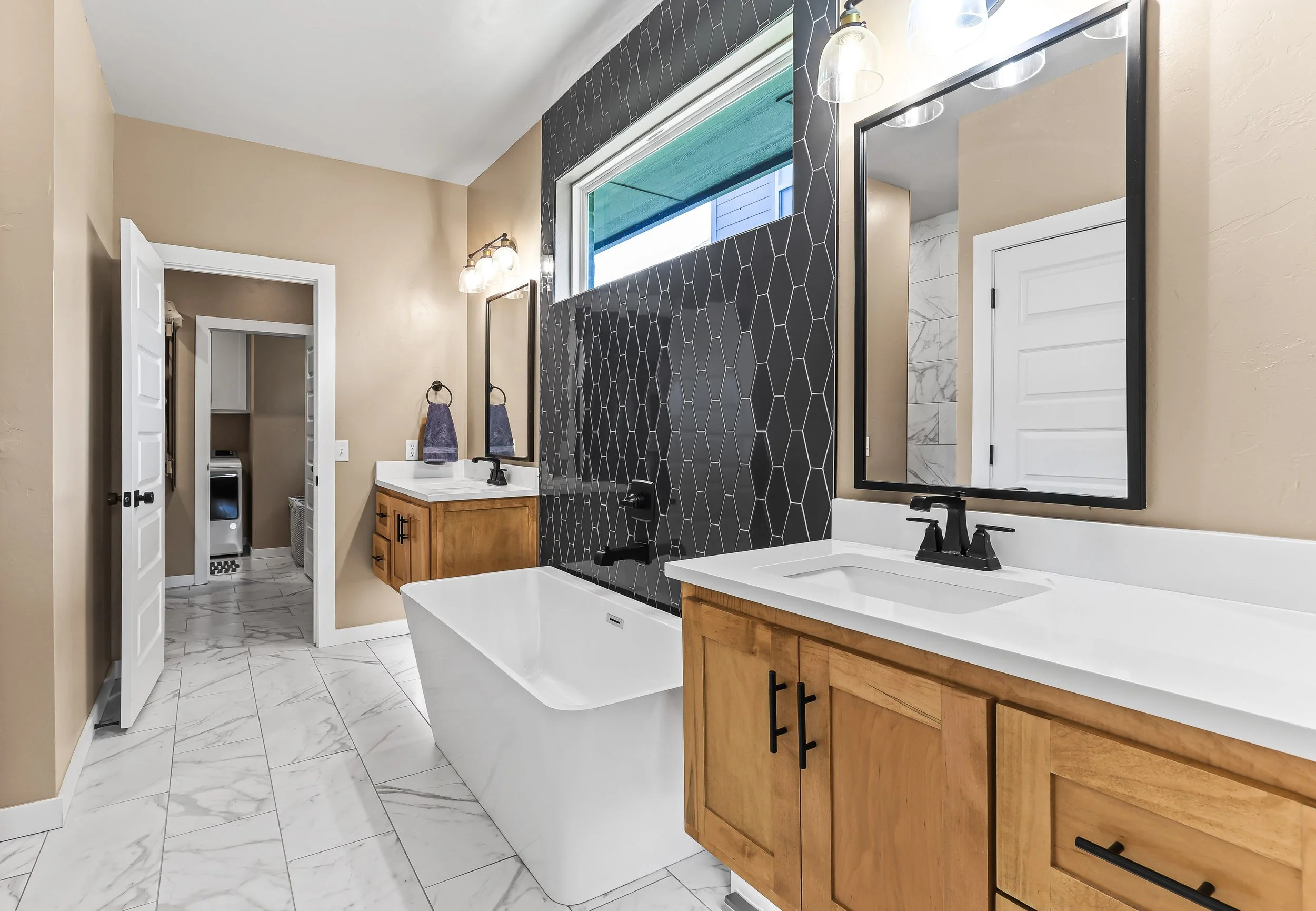 Modern bathroom with a white bathtub, wooden vanities, large mirrors, black fixtures, tiled walls, and a window with outdoor view. Real estate photography in Yukon, OK.