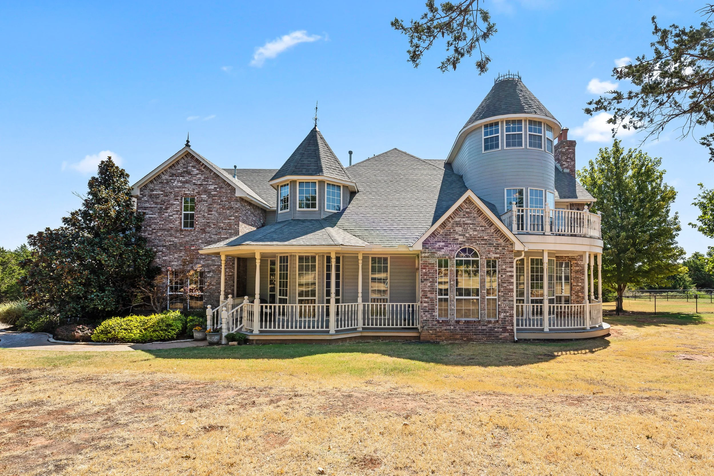 A large house with brick and gray siding, multiple gabled roofs, and a turret, surrounded by trees and shrubs, under a bright blue sky. Real estate photography in Mustang, OK.