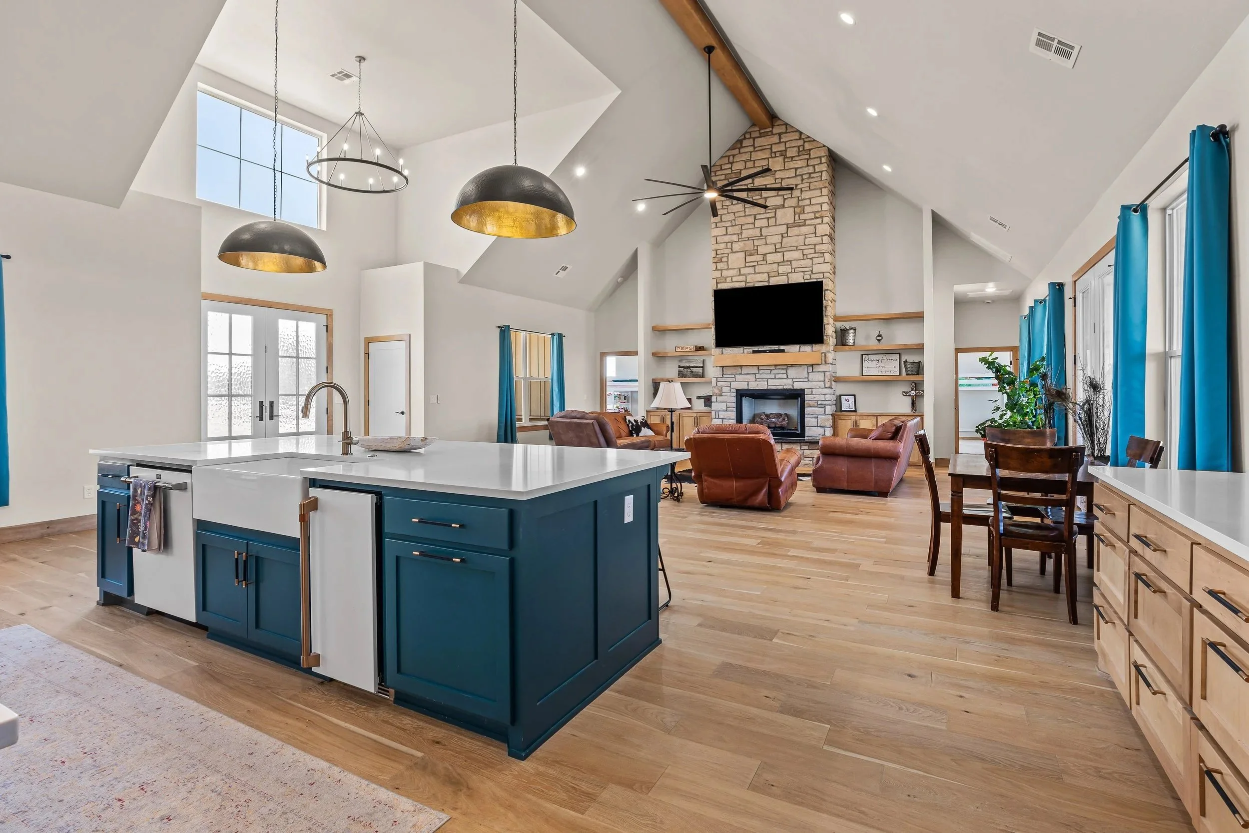 Open-concept living room and kitchen with vaulted ceiling, brick fireplace, large flat-screen TV, blue kitchen island, brown leather chairs, and large windows with blue curtains. Real estate photography in Geary, OK.