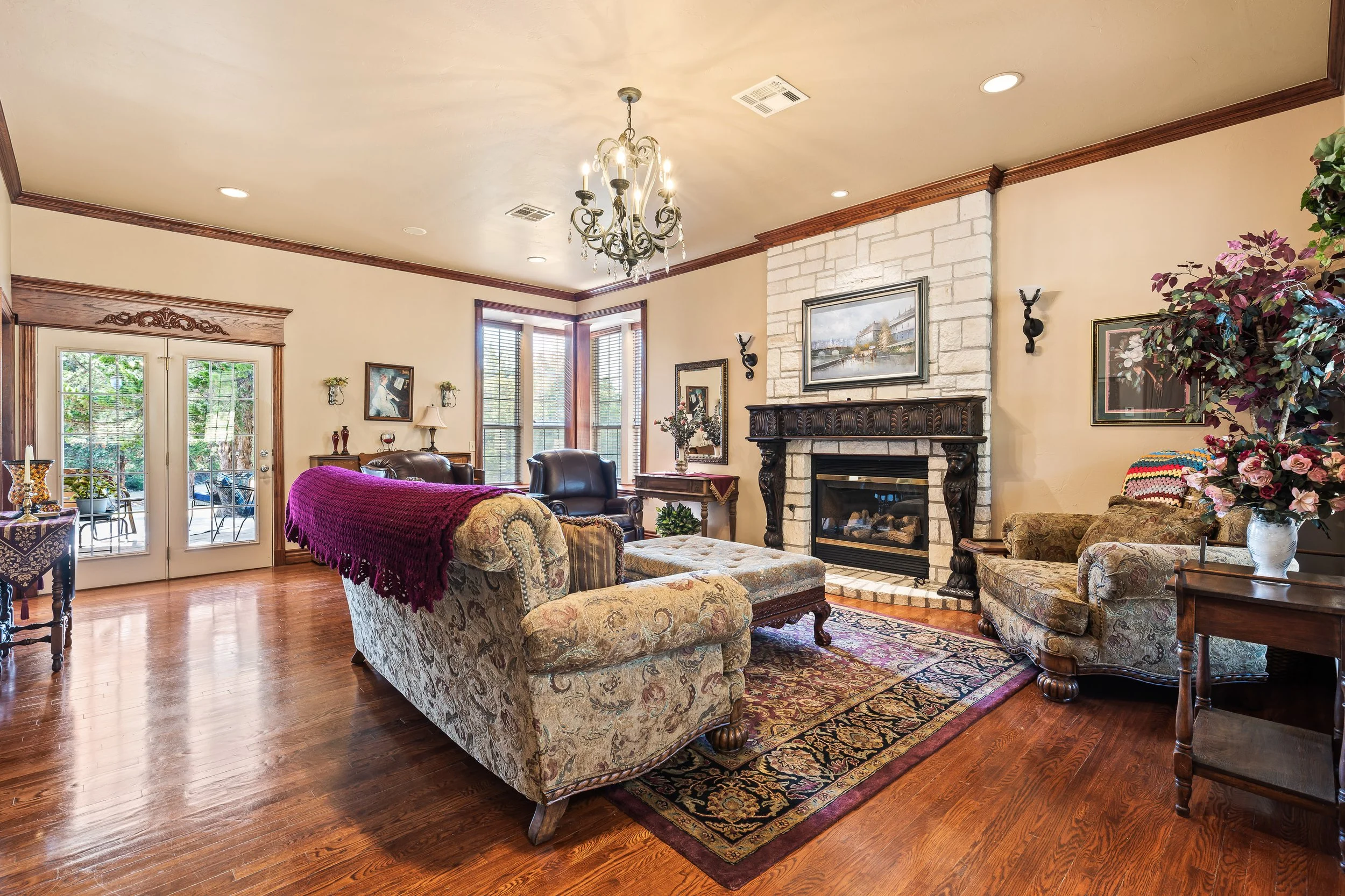 A spacious living room with hardwood floors, a stone fireplace with a wooden mantel, multiple sofas and chairs with floral and patterned upholstery, a chandelier hanging from the ceiling, paintings on the walls, a large plant, and a set of windows an