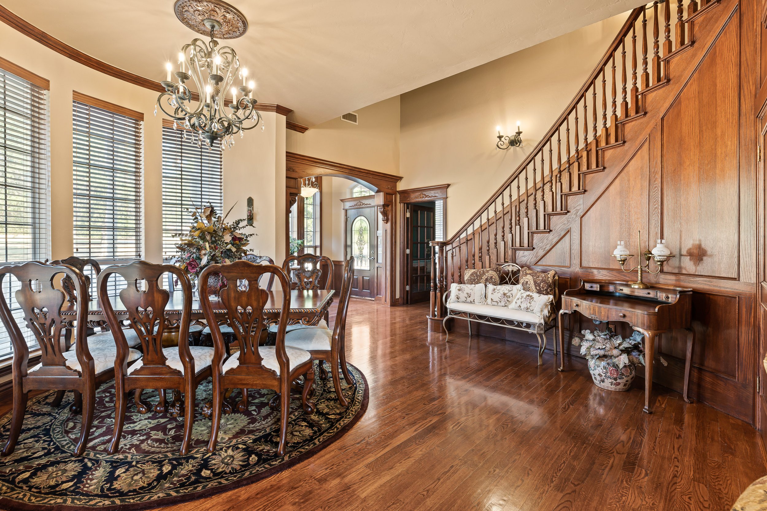 A spacious, well-lit dining room with a wooden dining table, matching chairs, a chandelier, large windows with blinds, a floral rug, and a staircase with wooden railings leading to an upper floor. The room features wood paneling and a polished hardwo
