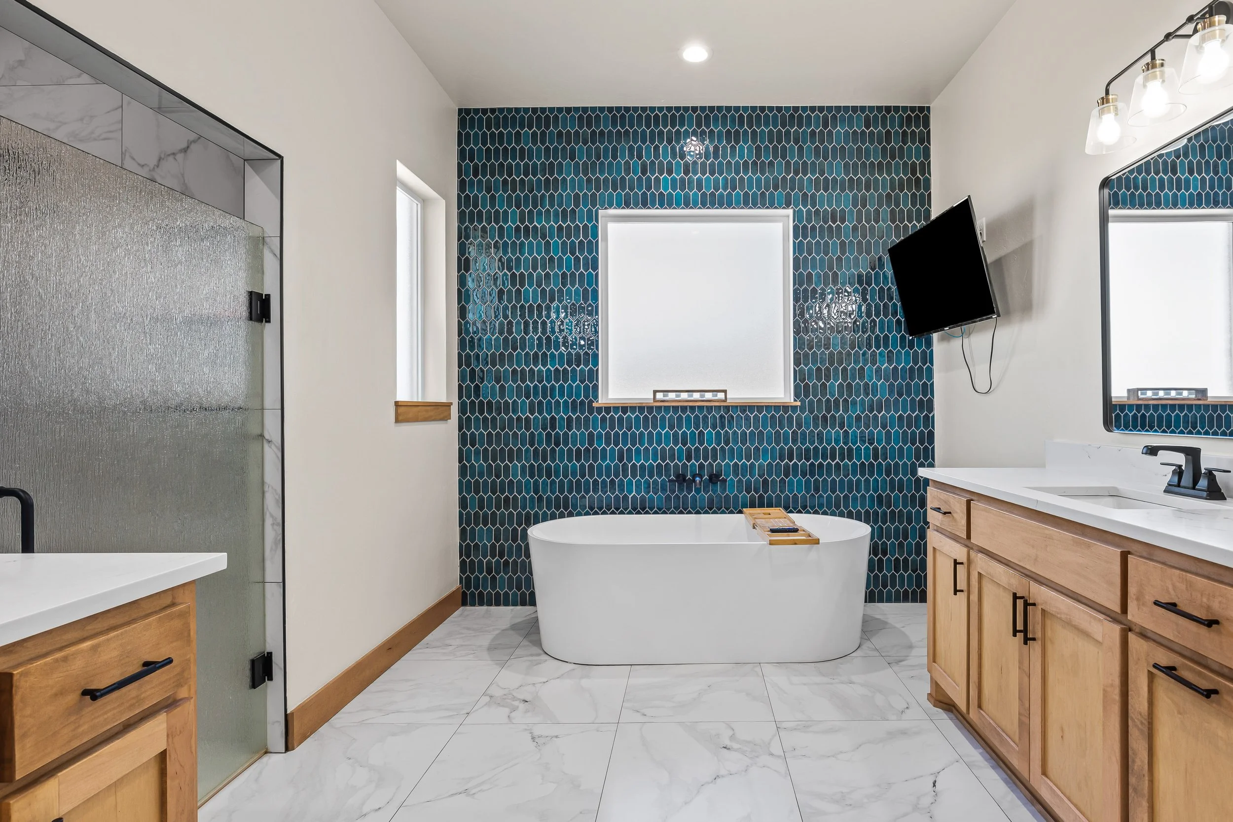 Modern bathroom with blue hexagonal tile accent wall, white freestanding bathtub, wooden vanity with white countertop, mounted flat-screen TV, and marble-style tile flooring. Real estate photography in Geary, OK.
