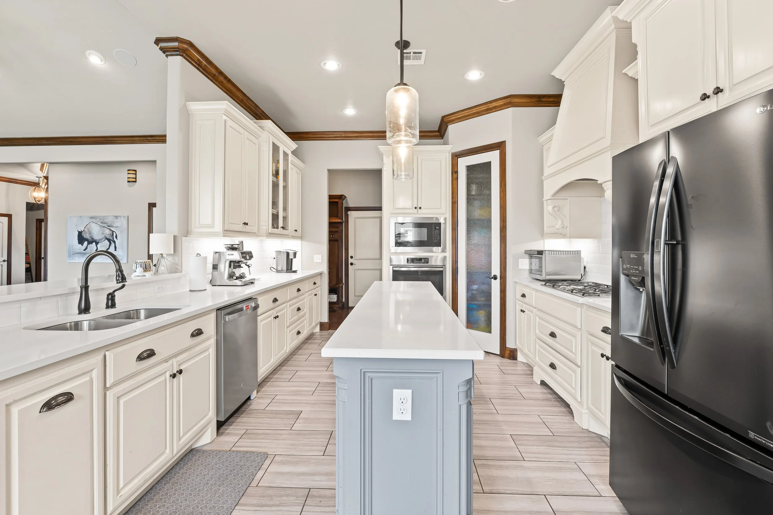 Modern white kitchen with island, stainless steel appliances, pendant lights, and wood trim ceiling. Real estate photography in Yukon, OK.