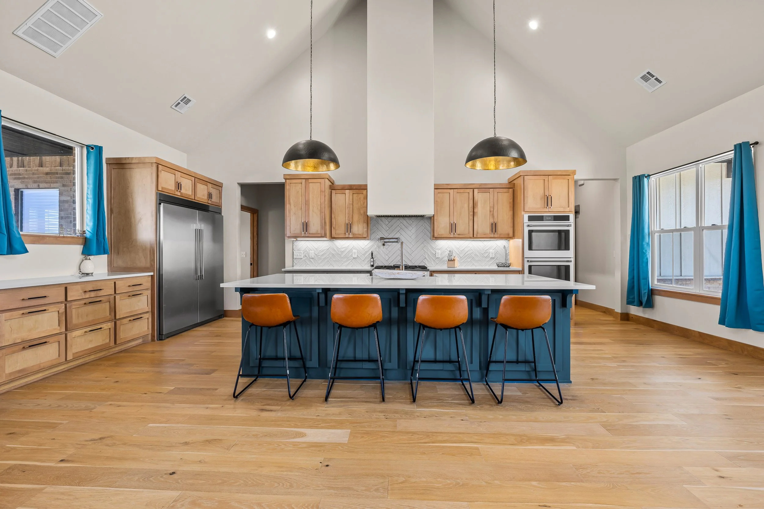 Kitchen with wooden cabinets, stainless steel refrigerator, white countertop, and a large island with blue base and orange chairs. Blue curtains and large windows with natural light. Wooden floor and modern lighting fixtures. Real estate photography 