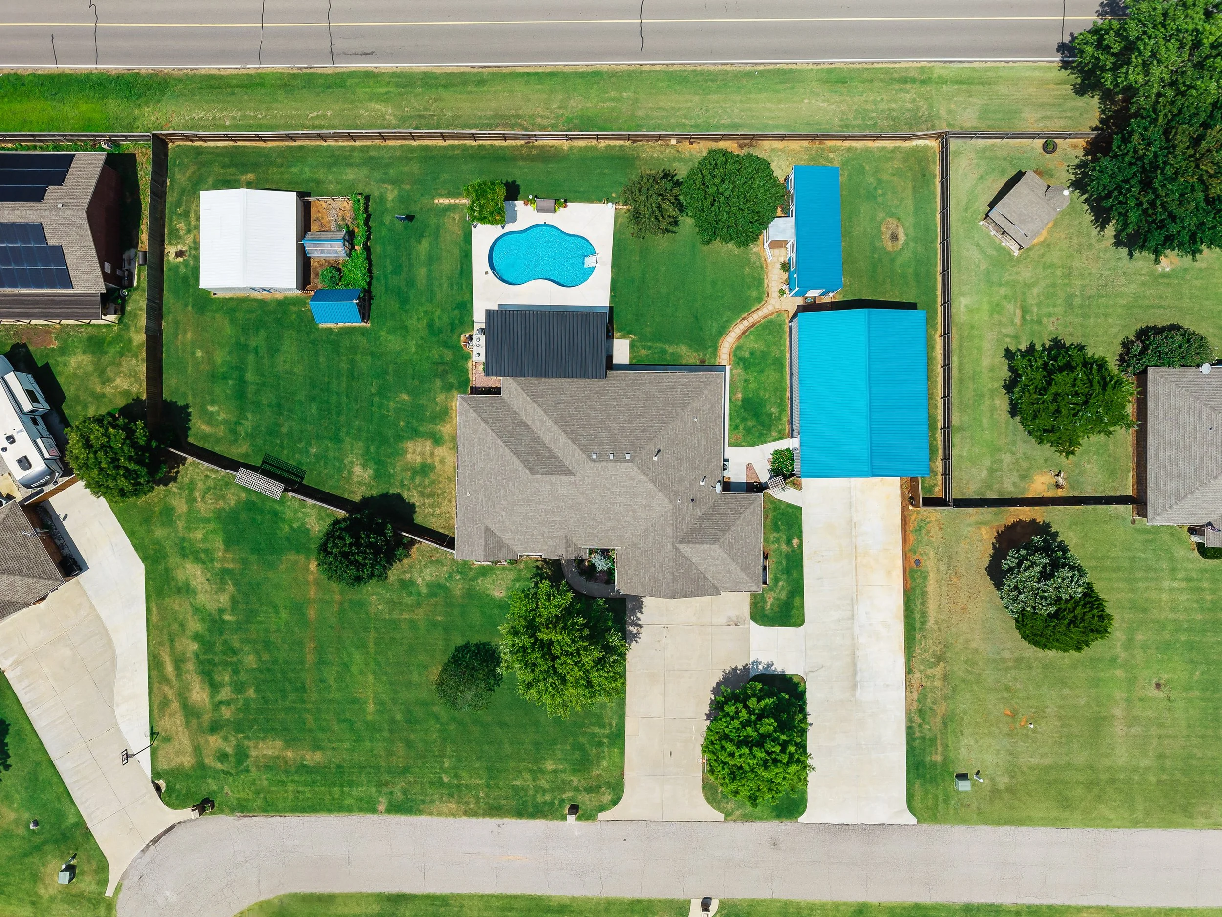 An aerial view of a suburban backyard featuring a kidney-shaped swimming pool with a concrete deck, surrounded by green grass, trees, and two small blue sheds. There is a house with a gray roof and adjacent driveway with a white vehicle. A sidewalk b