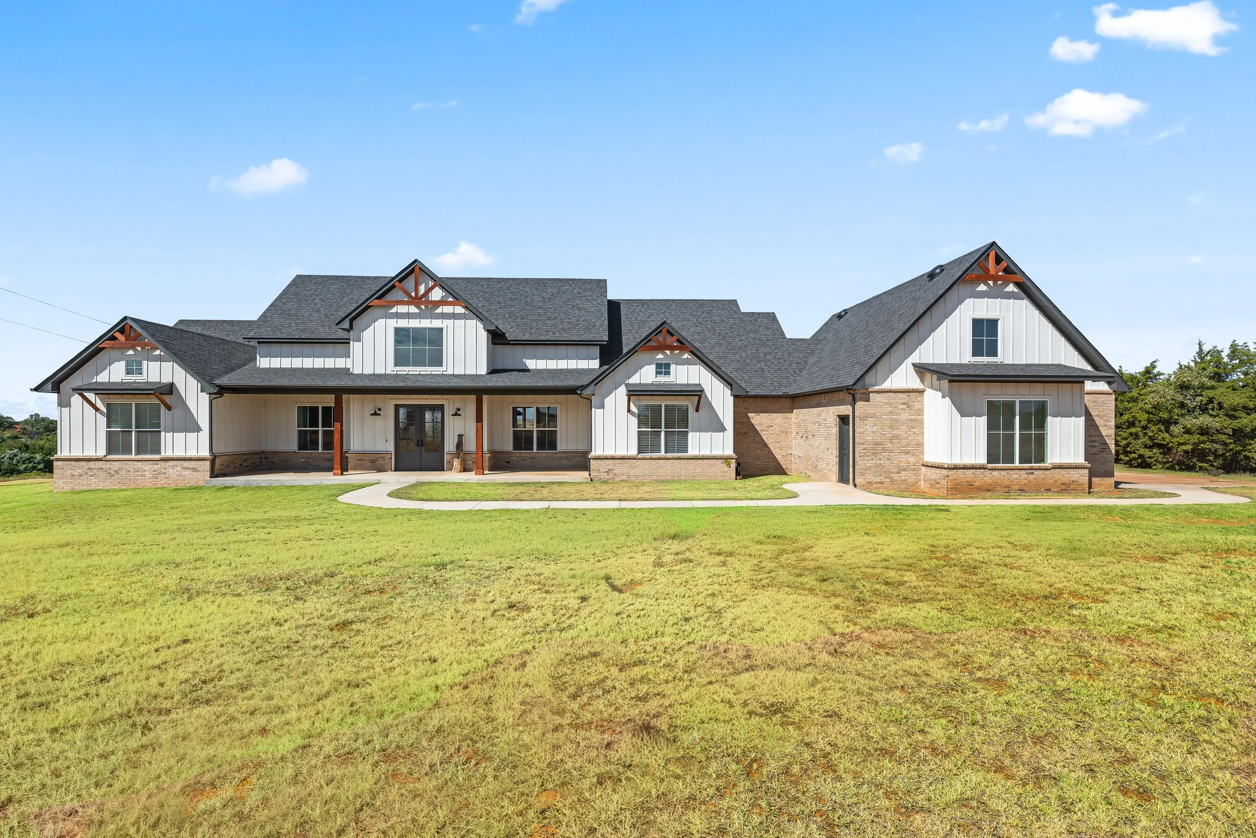 A large house with a mix of farm-style and modern architecture, featuring a dark gray roof, white vertical siding, and brick accents, situated on a well-kept grassy yard under a partly cloudy blue sky. Real estate photography in Geary, OK.