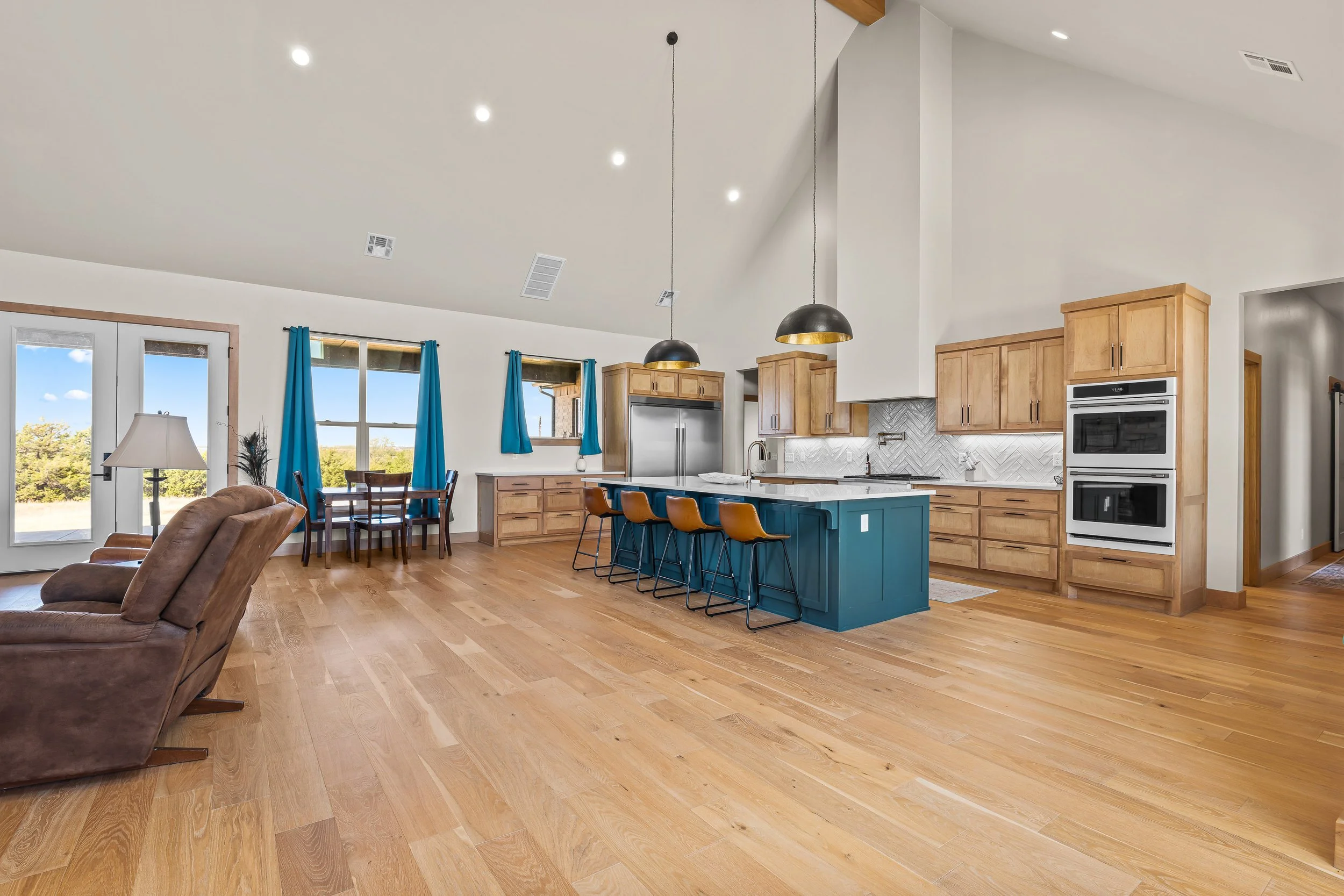 Open-concept kitchen and living room with hardwood floors, white walls, natural wood cabinets, a blue kitchen island, black pendant lights, large windows with blue curtains, and a patio door. Real estate photography in Geary, OK.