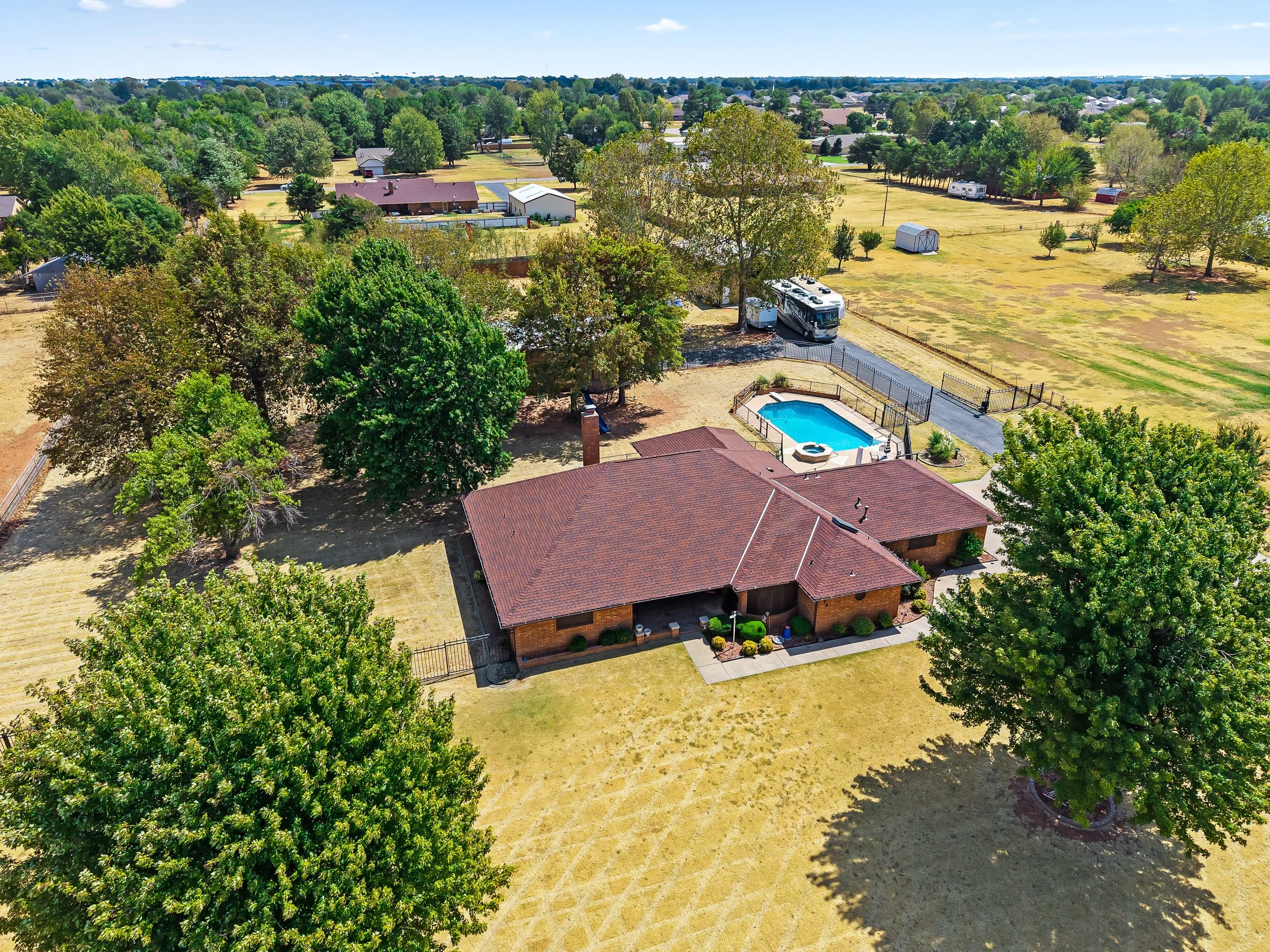 Aerial view of a suburban backyard with a swimming pool, trees, a house with a red roof, and surrounding neighborhood with houses and green trees. Drone Photography in Mustang OK.