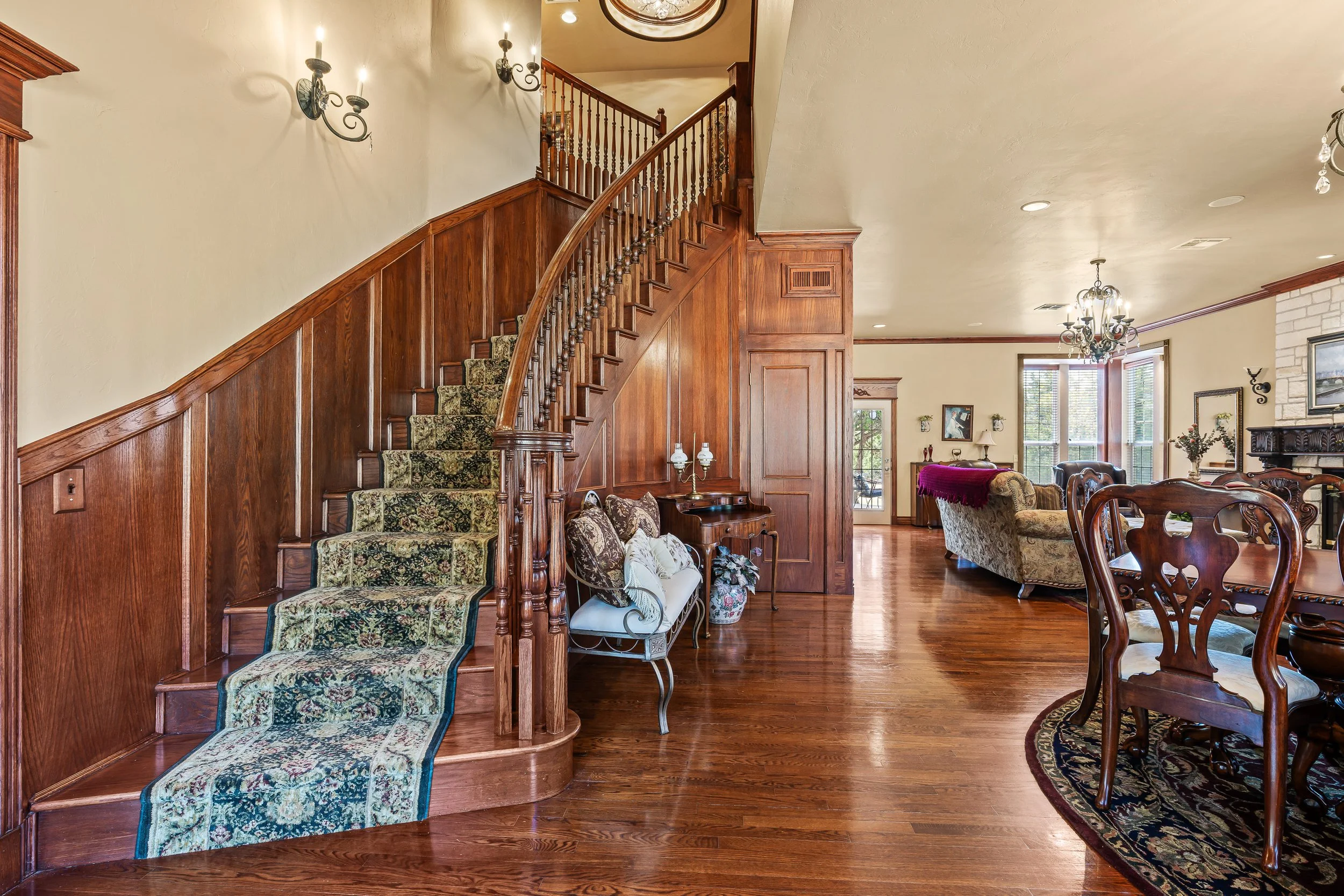 Interior view of a living room with wooden staircase, floral carpet runners on stairs, traditional furniture including sofas, armchairs, and dining table, chandelier, wall sconces, and brick fireplace. Real estate photography in Mustang, OK.