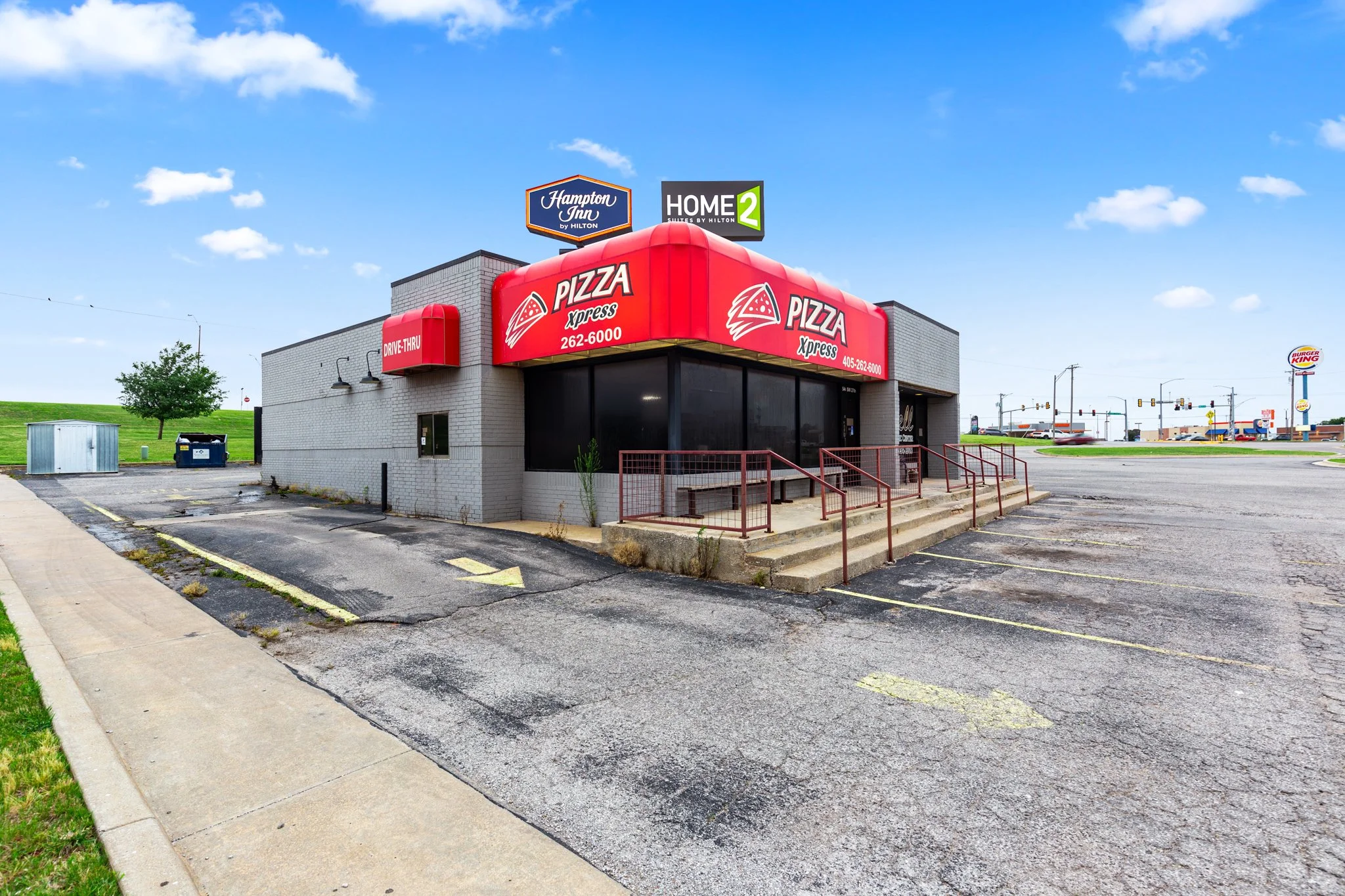 A small fast food restaurant building with a large red Pizza Xpress sign on the front. The building is gray brick, with a staircase leading to the entrance, and has drive-thru signage. In the background, there are signs for Hampton Inn and Home2 Suit