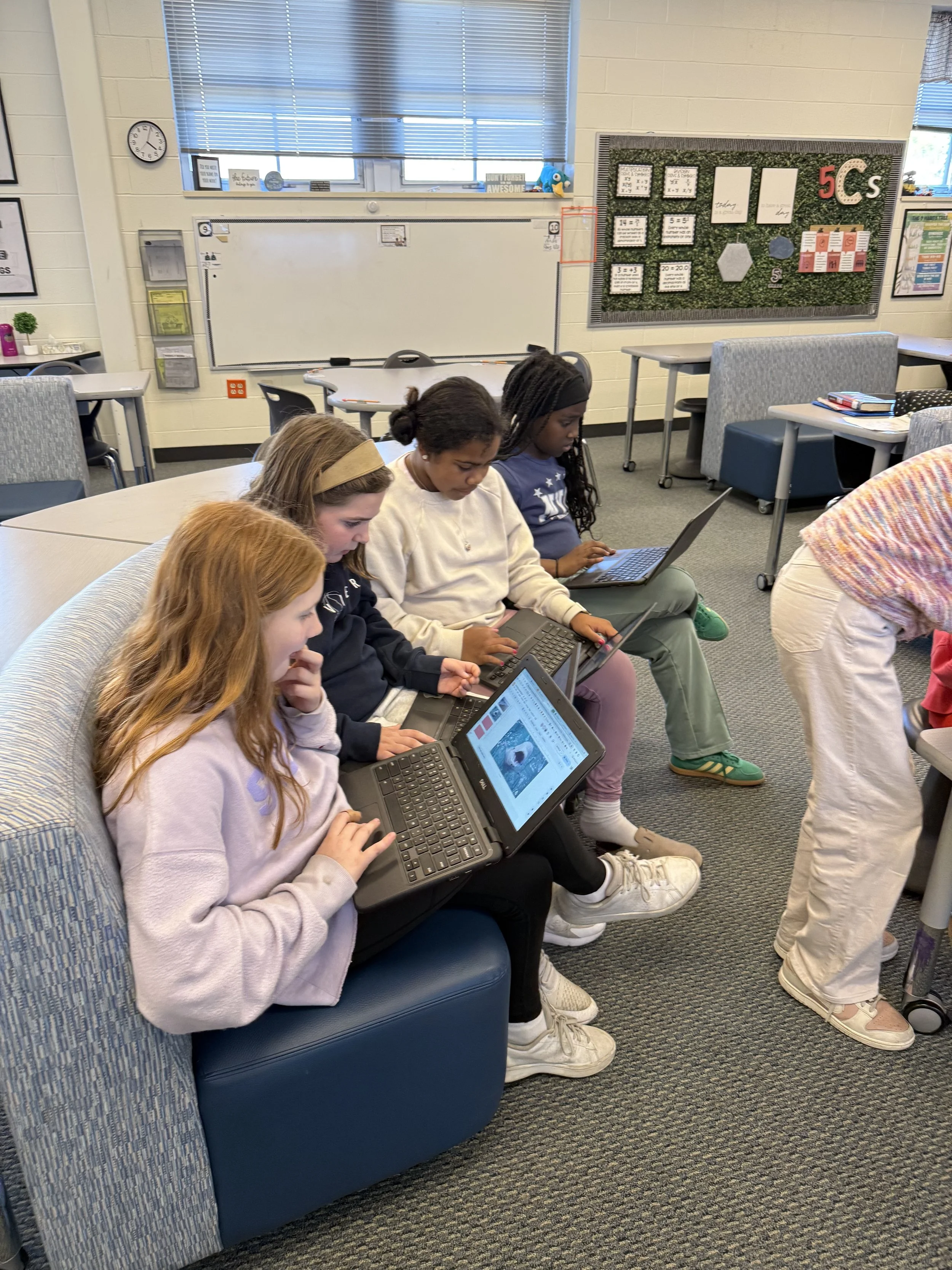 Four students sitting on a couch in a classroom, each working on a laptop, with a teacher bending over in front of them.