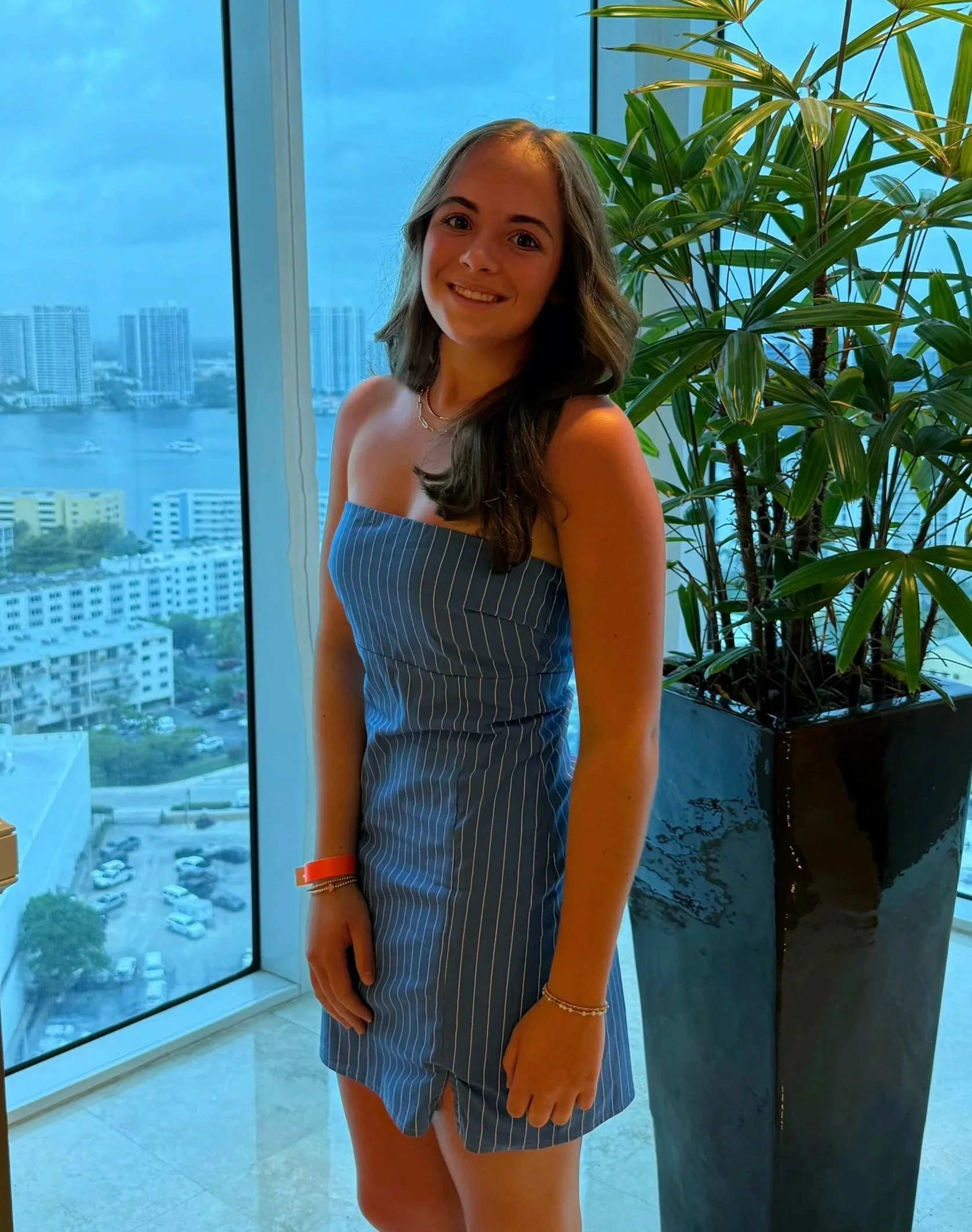 Young woman standing near window with a city skyline and water in the background, wearing a striped blue dress and smiling.