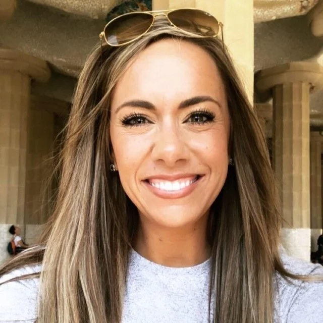A woman smiling, with long brown hair, wearing sunglasses on her head and a light-colored top.