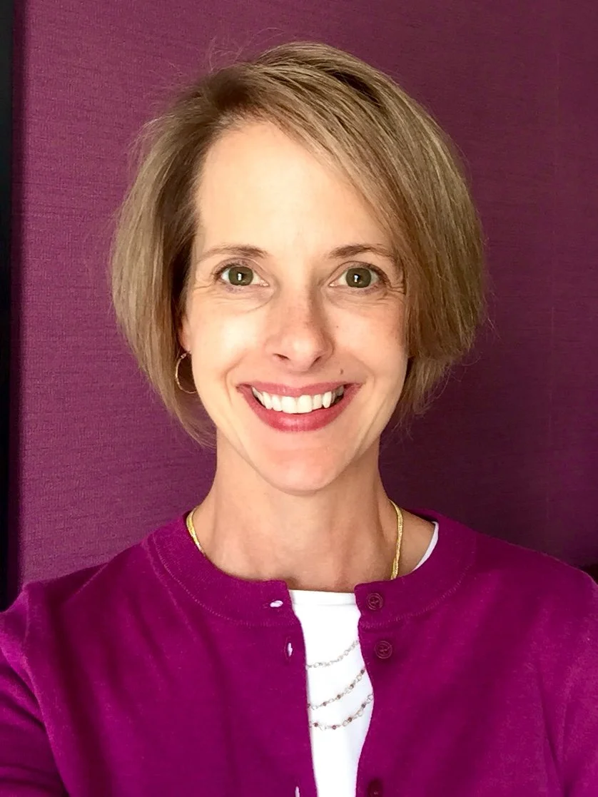 A woman with short blonde hair, smiling, wearing a purple cardigan over a white shirt, gold necklace, and hoop earrings, standing against a purple background.