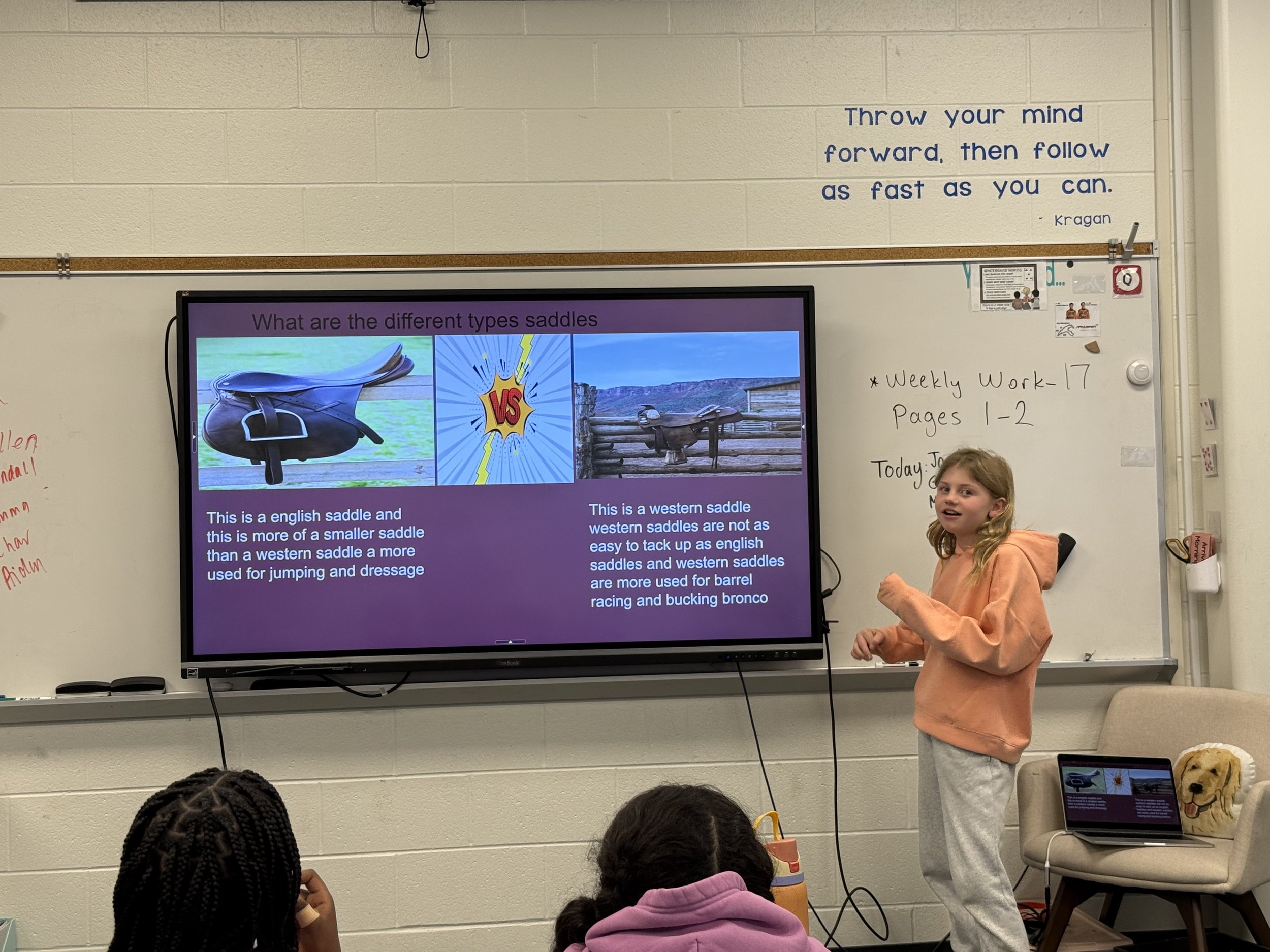 A classroom presentation featuring a girl in an orange hoodie and gray pants standing next to a large screen. The screen displays a comparison of English and western saddles with images and descriptions. Several students sit facing the screen, and a 