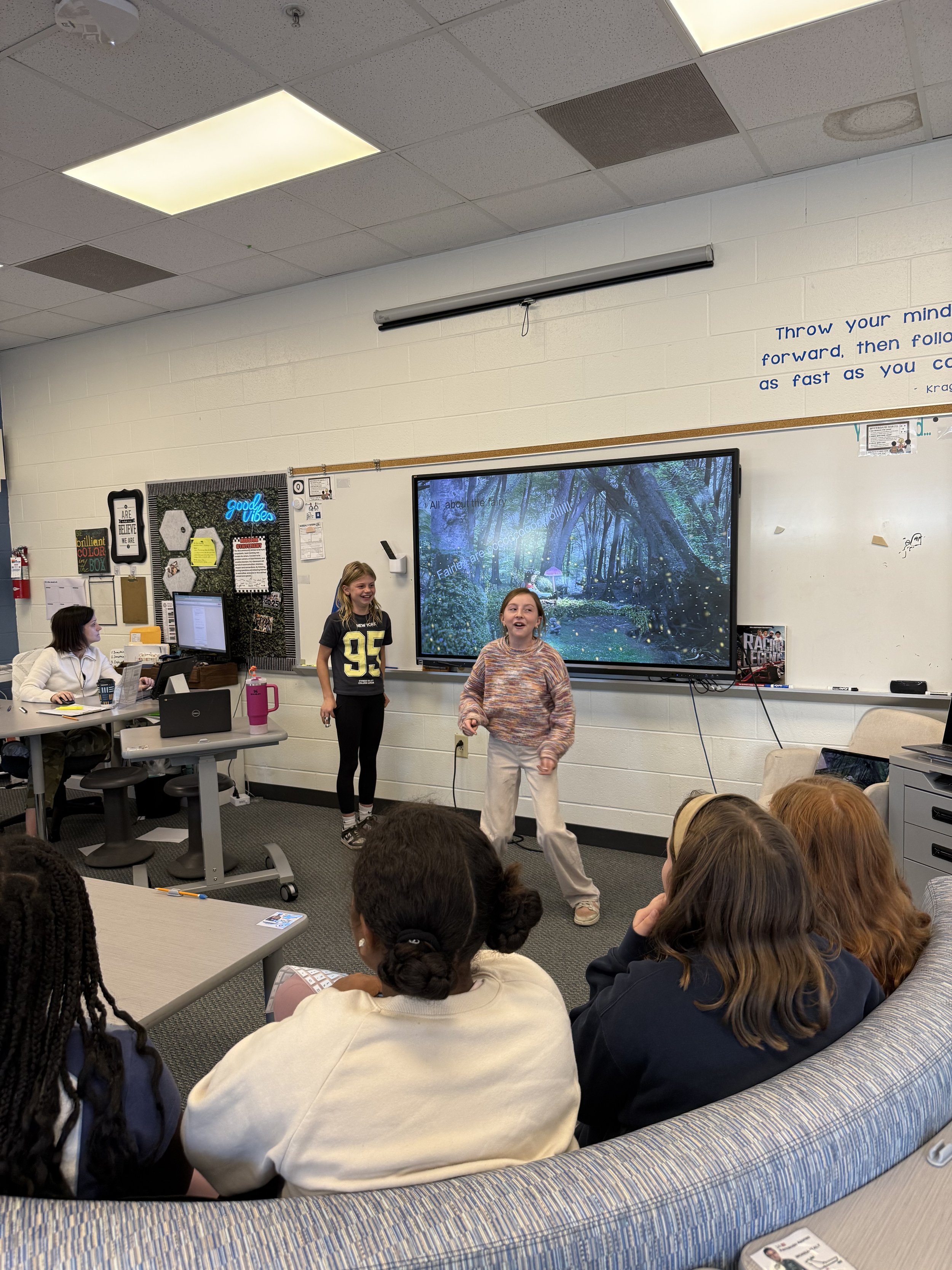 A classroom with students and teachers. Two girls are presenting at the front, one standing and the other sitting. Several students are sitting on a curved sofa, watching the presentation. The classroom has a large screen displaying a forest scene, a
