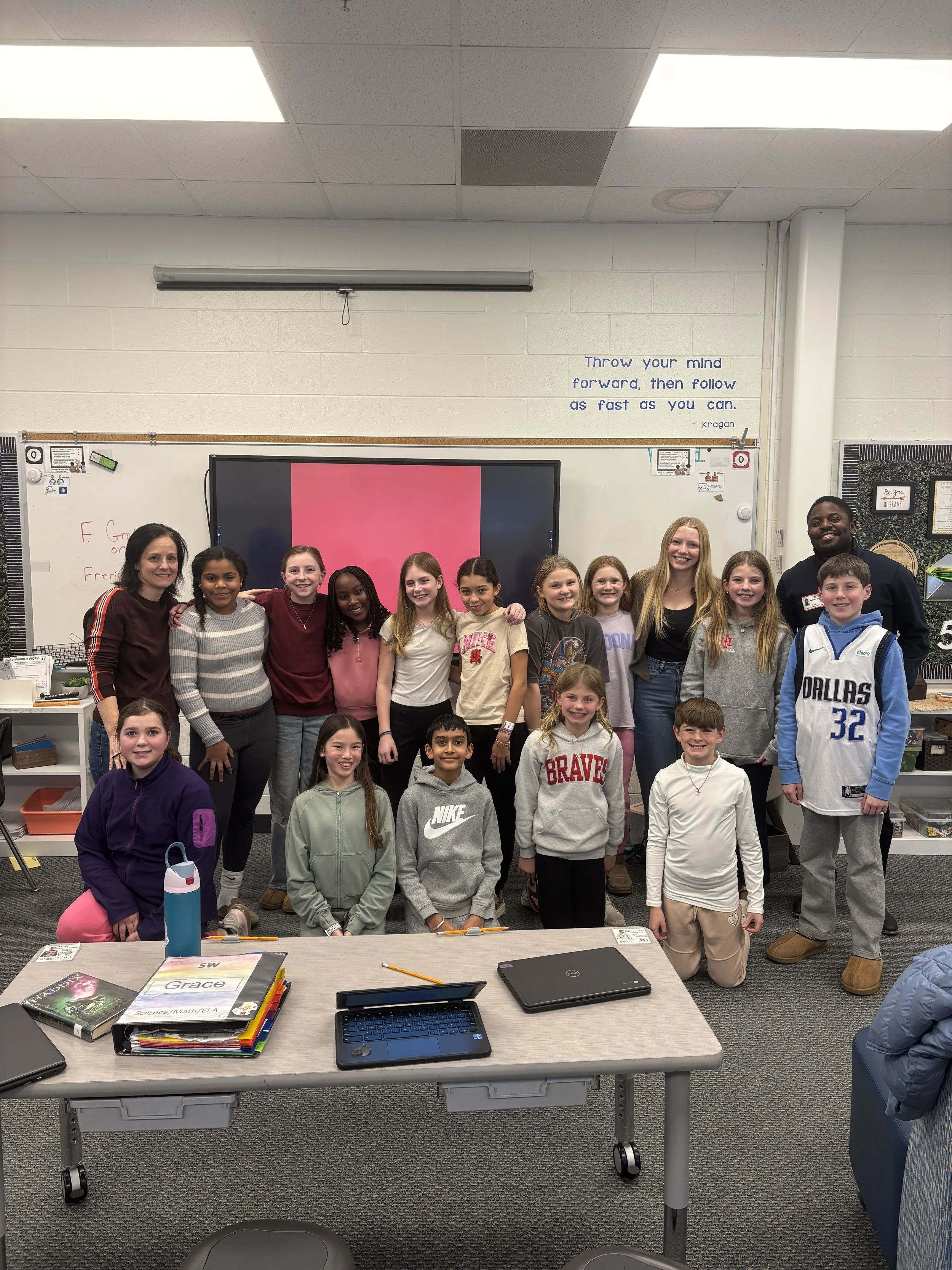 A classroom group photo of children with two teachers, smiling and standing in front of a whiteboard and a large screen. The classroom has desks with books, laptops, and school supplies. The whiteboard has some writings, and a quote is posted on the 