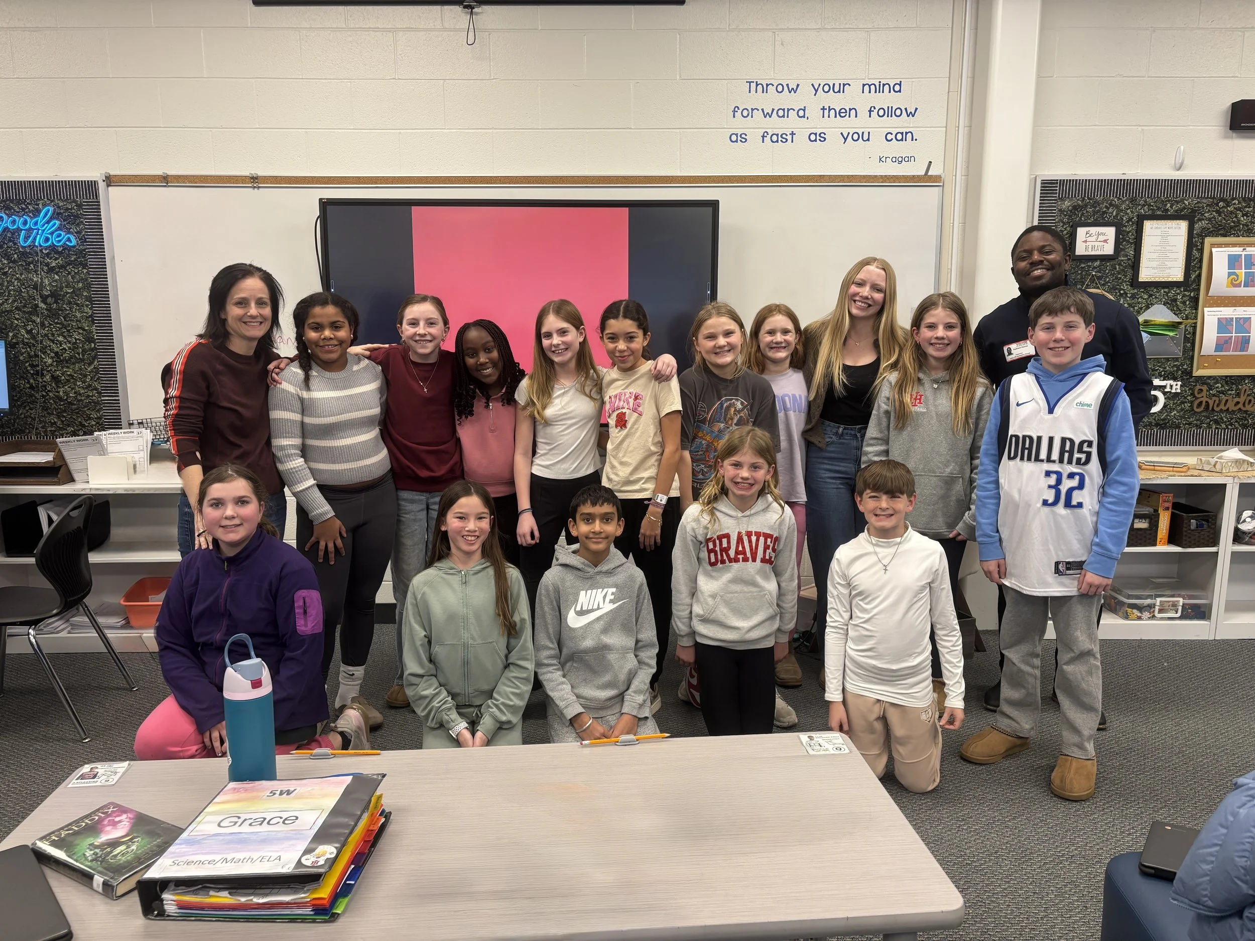 Group photo of young students and two teachers in a classroom, smiling at the camera, with a large screen and educational posters in the background.