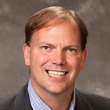 A man with brown hair smiling, wearing a suit jacket over a blue checkered shirt, against a gray background.