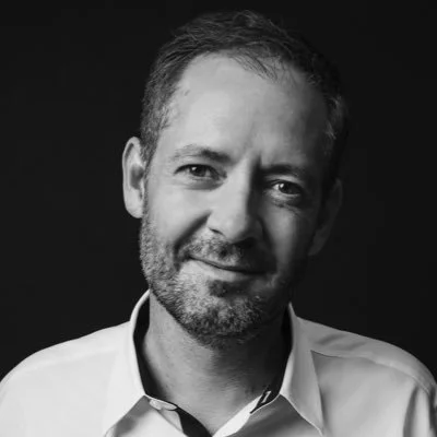Black and white portrait of a man with short hair and a beard, wearing a collared shirt, smiling slightly against a dark background.
