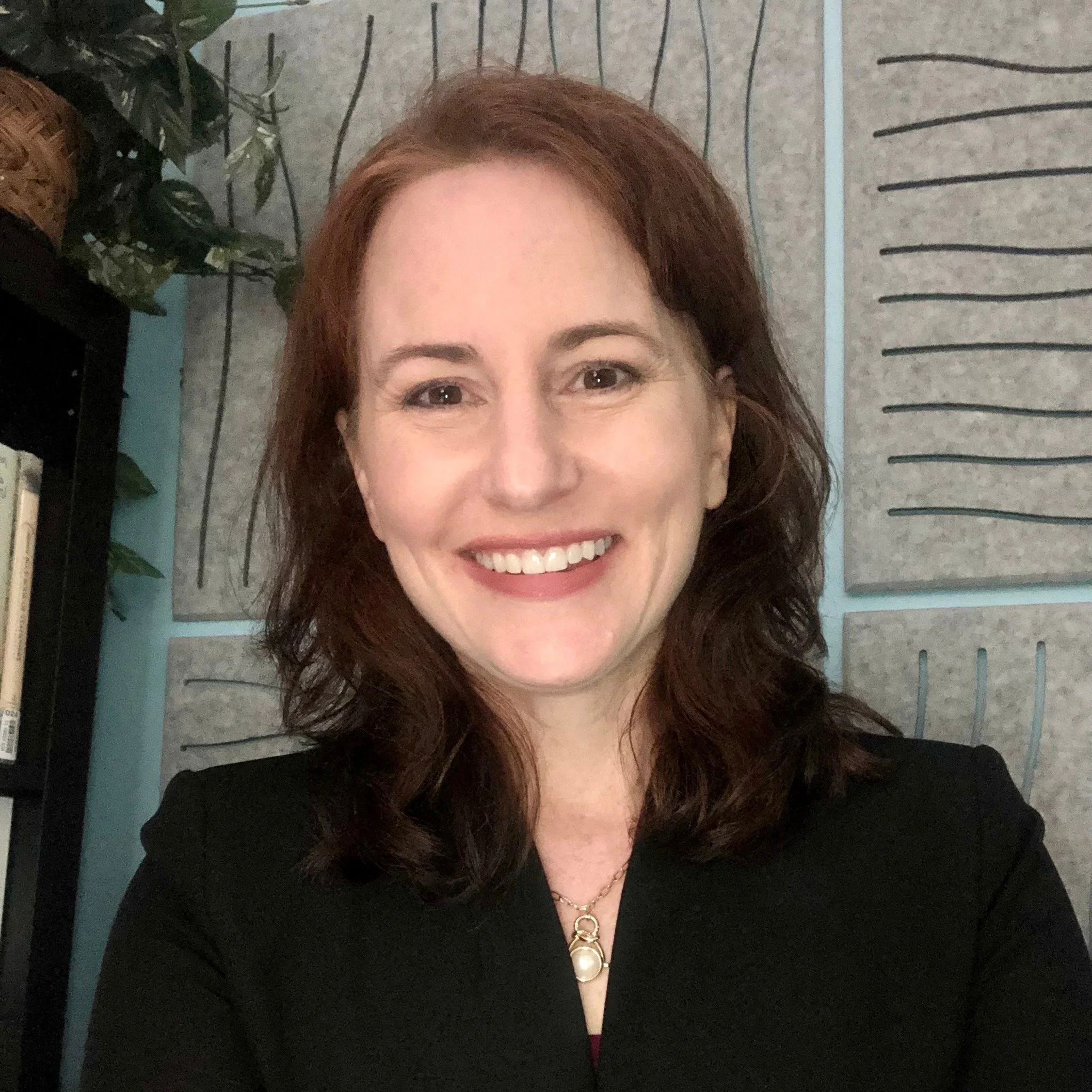 A woman with shoulder-length wavy auburn hair, smiling, wearing a black top and a pearl necklace. She is in a room with soundproofing panels and a bookshelf, and a plant is partially visible to her left.