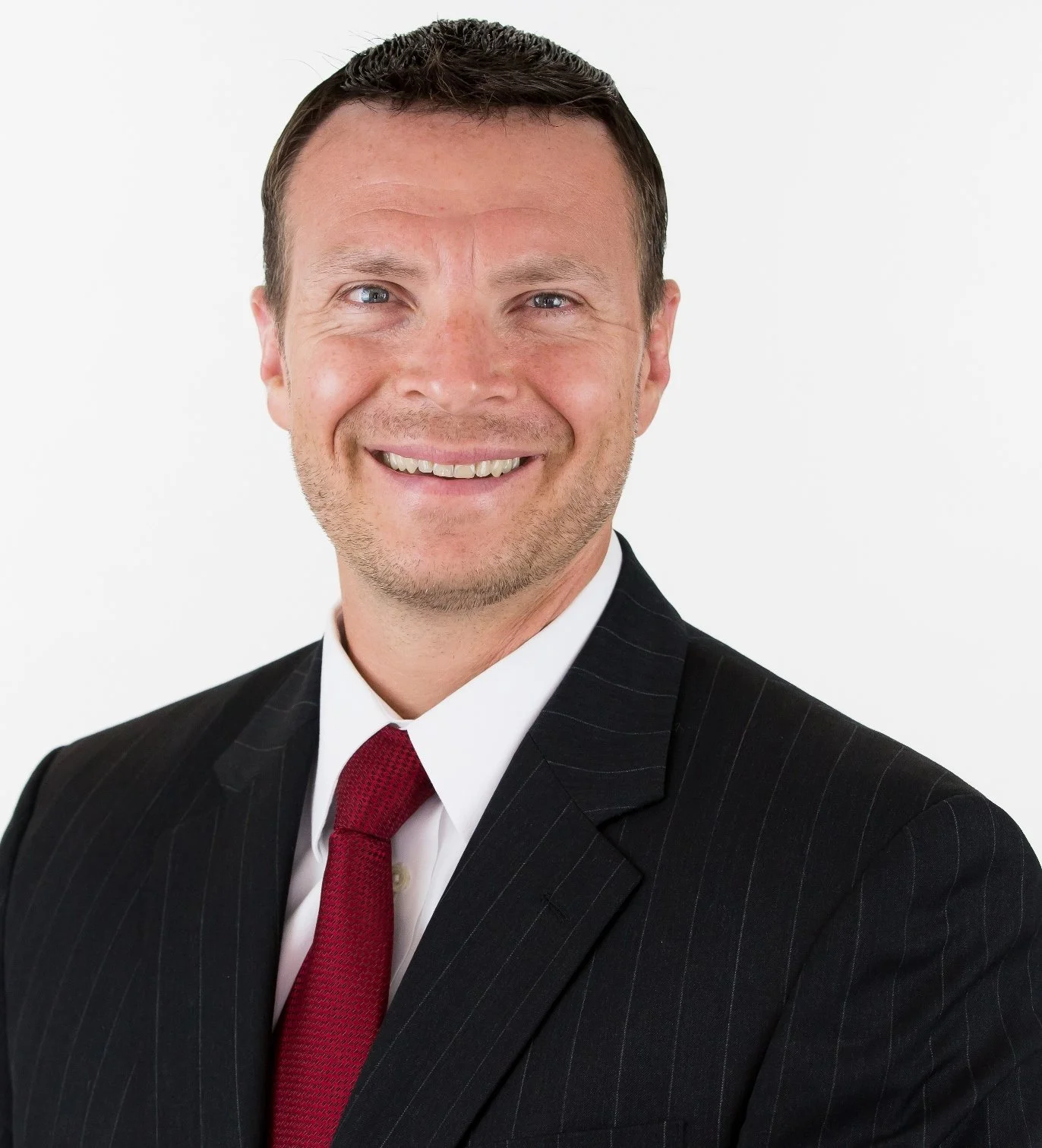 Professional headshot of a smiling man wearing a black pinstripe suit, white shirt, and red tie, against a white background.