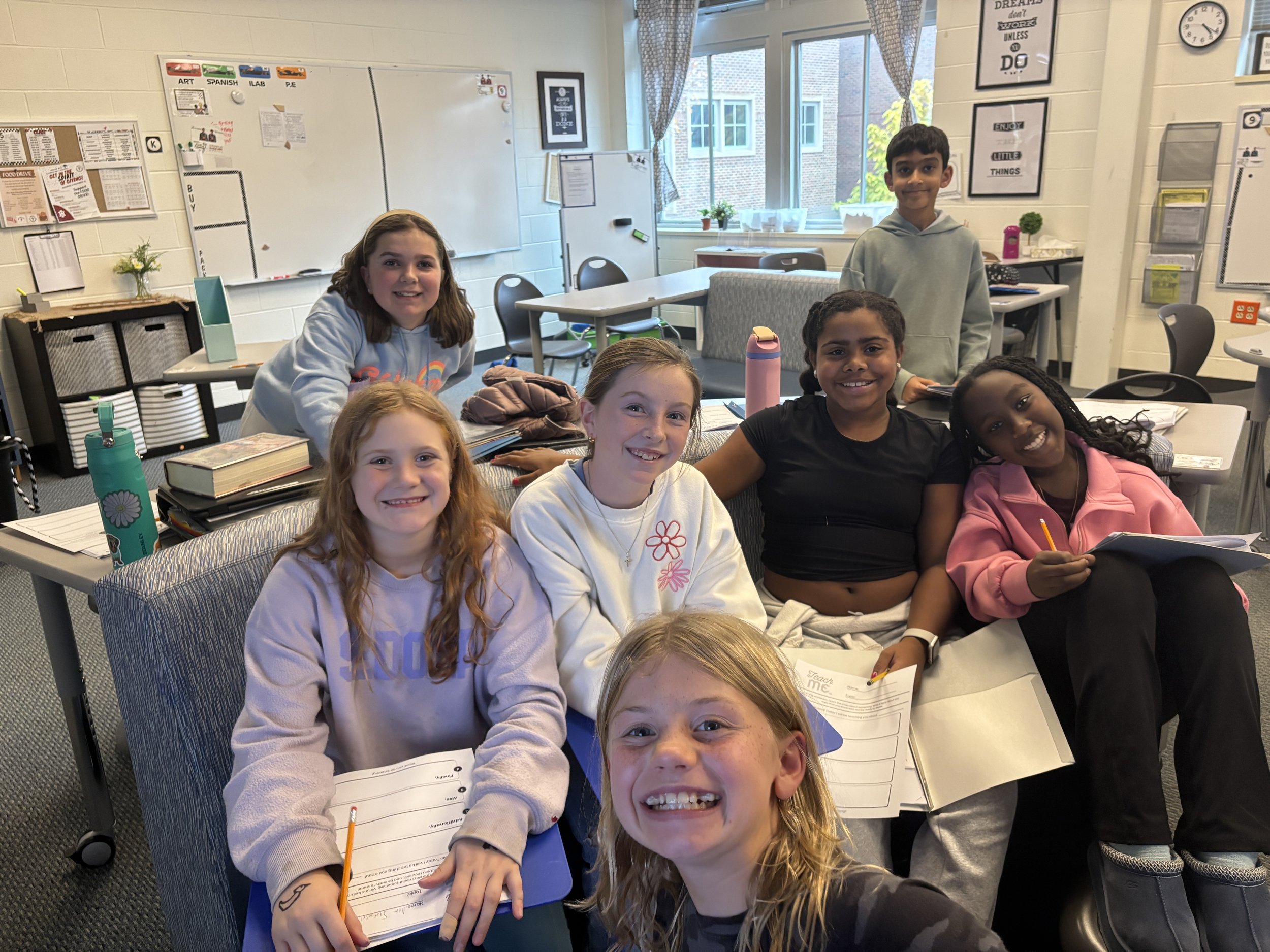 Group of seven children sitting around a table in a classroom, smiling at the camera. The classroom has educational posters, whiteboards, and windows with curtains in the background.