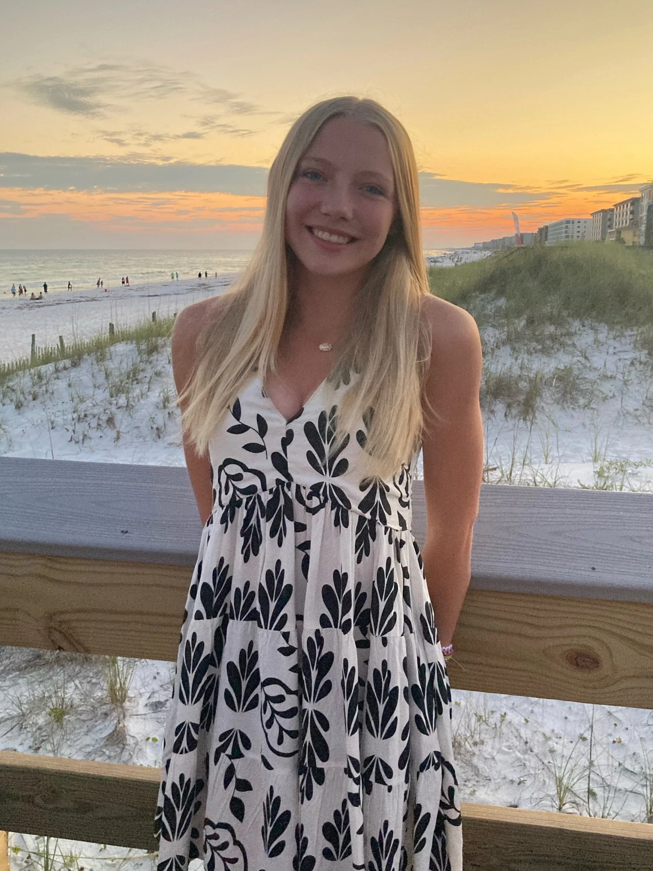 Young woman with long blonde hair smiling on a beach at sunset, wearing a sleeveless dress with a black and white pattern, standing behind a wooden railing.