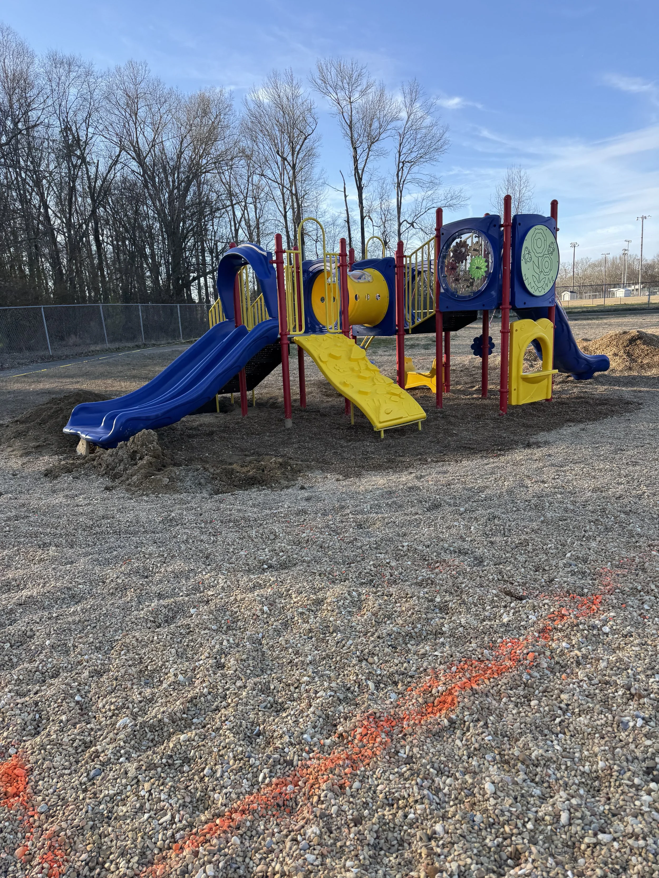Empty playground with slides, climbing equipment, and surrounding trees without leaves under a partly cloudy sky.