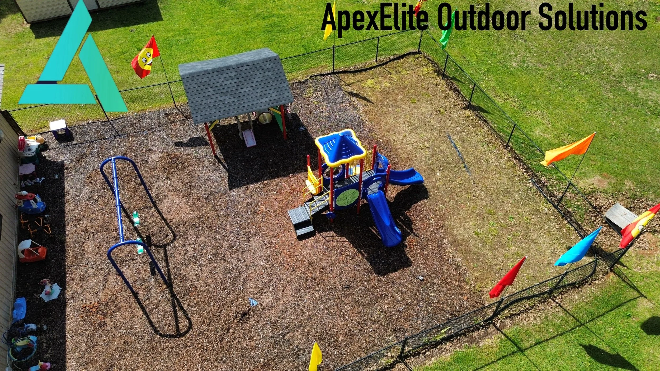 Child's playground with blue slide, small house, and various toys, surrounded by a black fence. Flag banners are visible, and part of a green lawn extends beyond the play area.