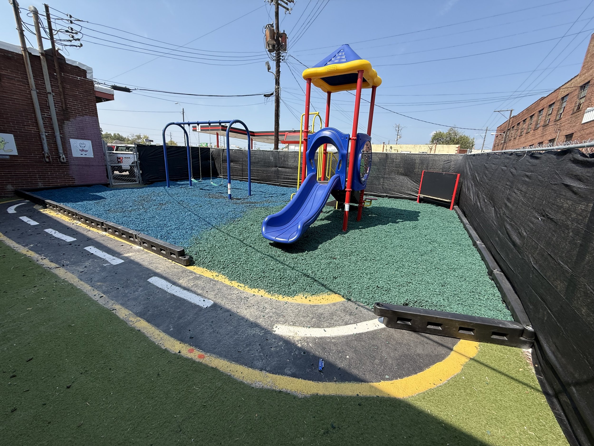 An empty playground with a blue slide, swings, and a small climbing wall, surrounded by black fencing and a brick building in the background, under a clear blue sky.
