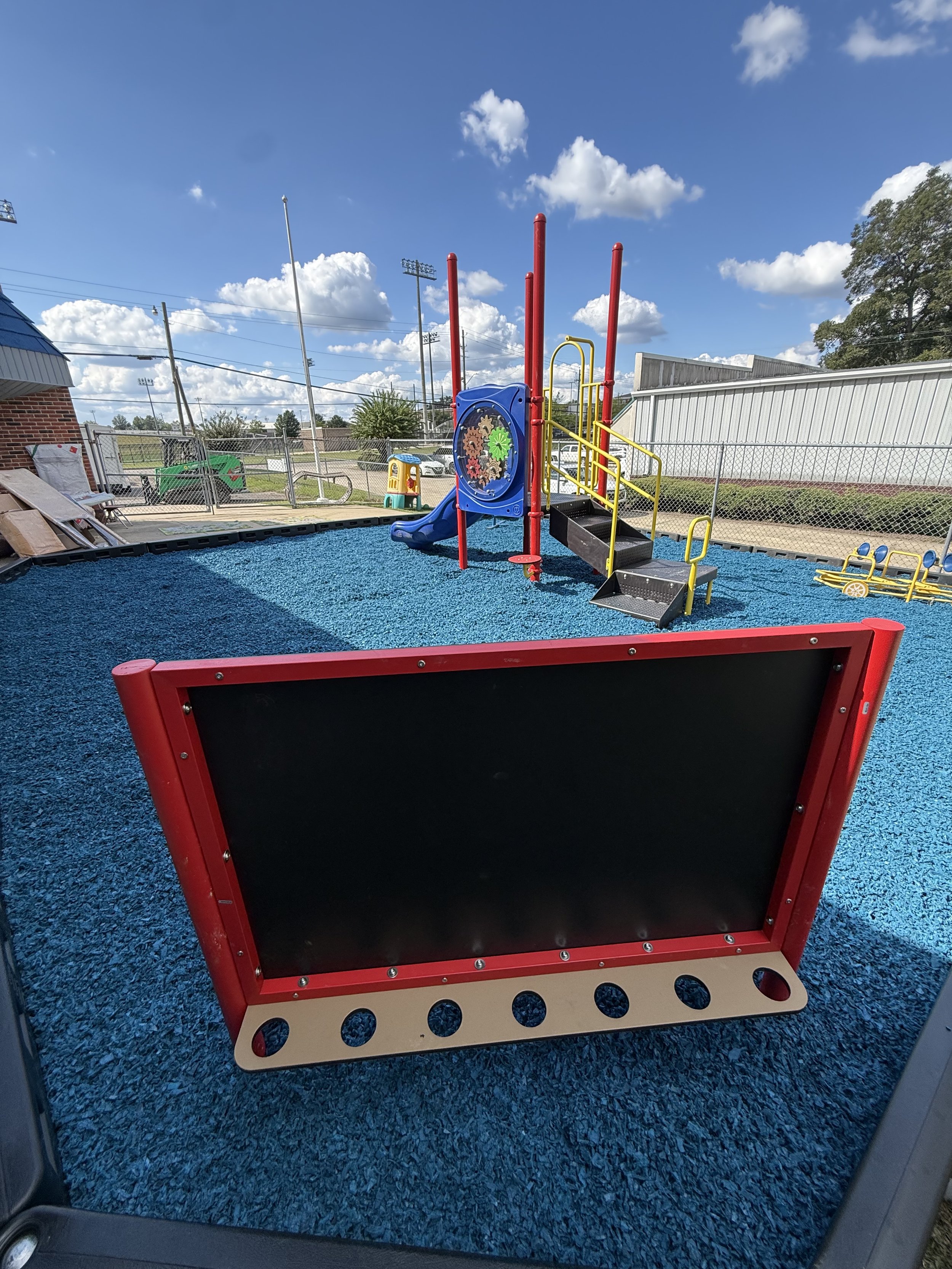 Playground with a blue rubber floor, and a playing structure with a small slide, climbing bars, and gear decorations, enclosed by a wire fence, under a blue sky with clouds.