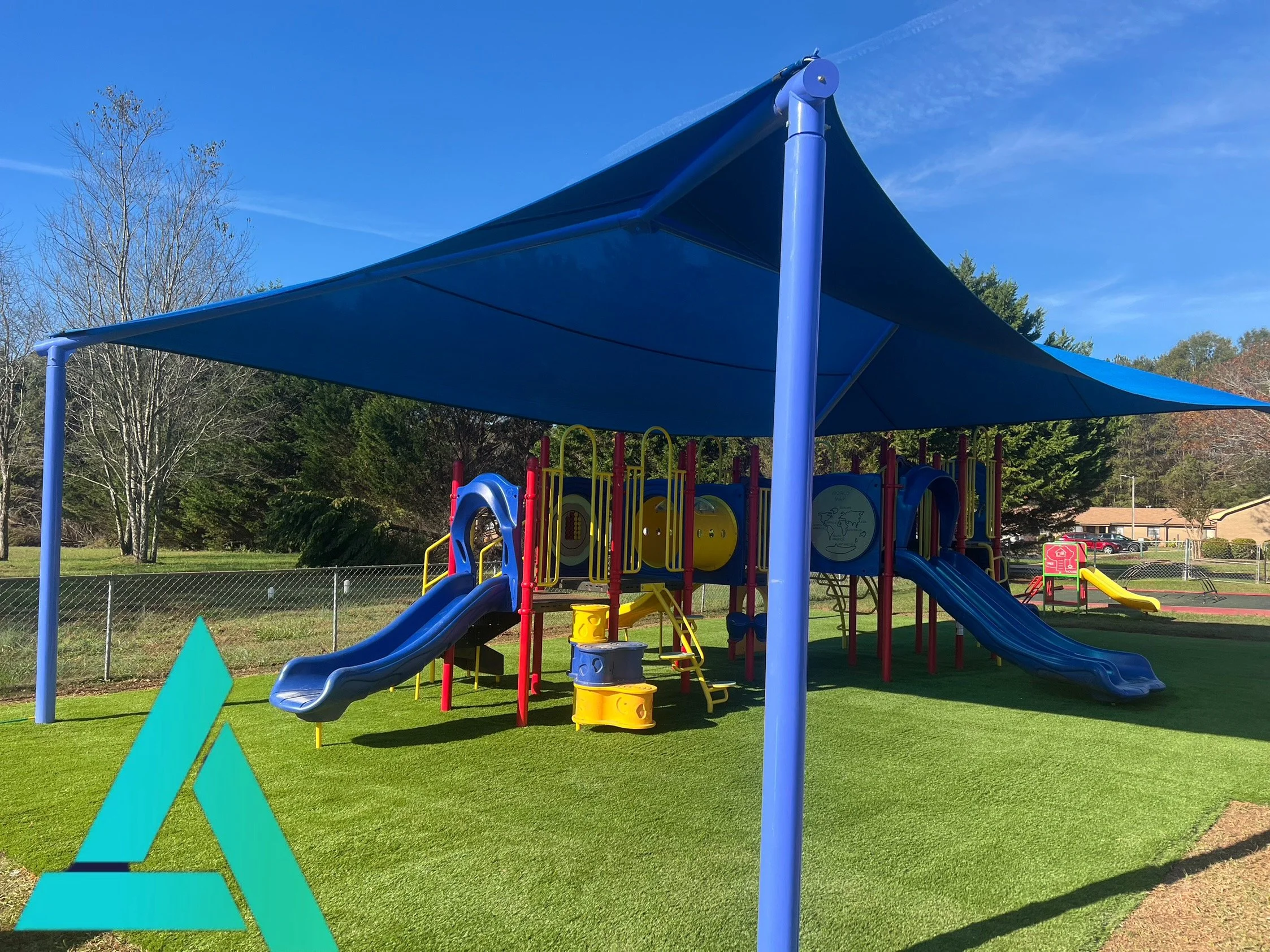 Colorful playground equipment under a blue sunshade in a park, surrounded by trees and a fence, with a clear sky above.