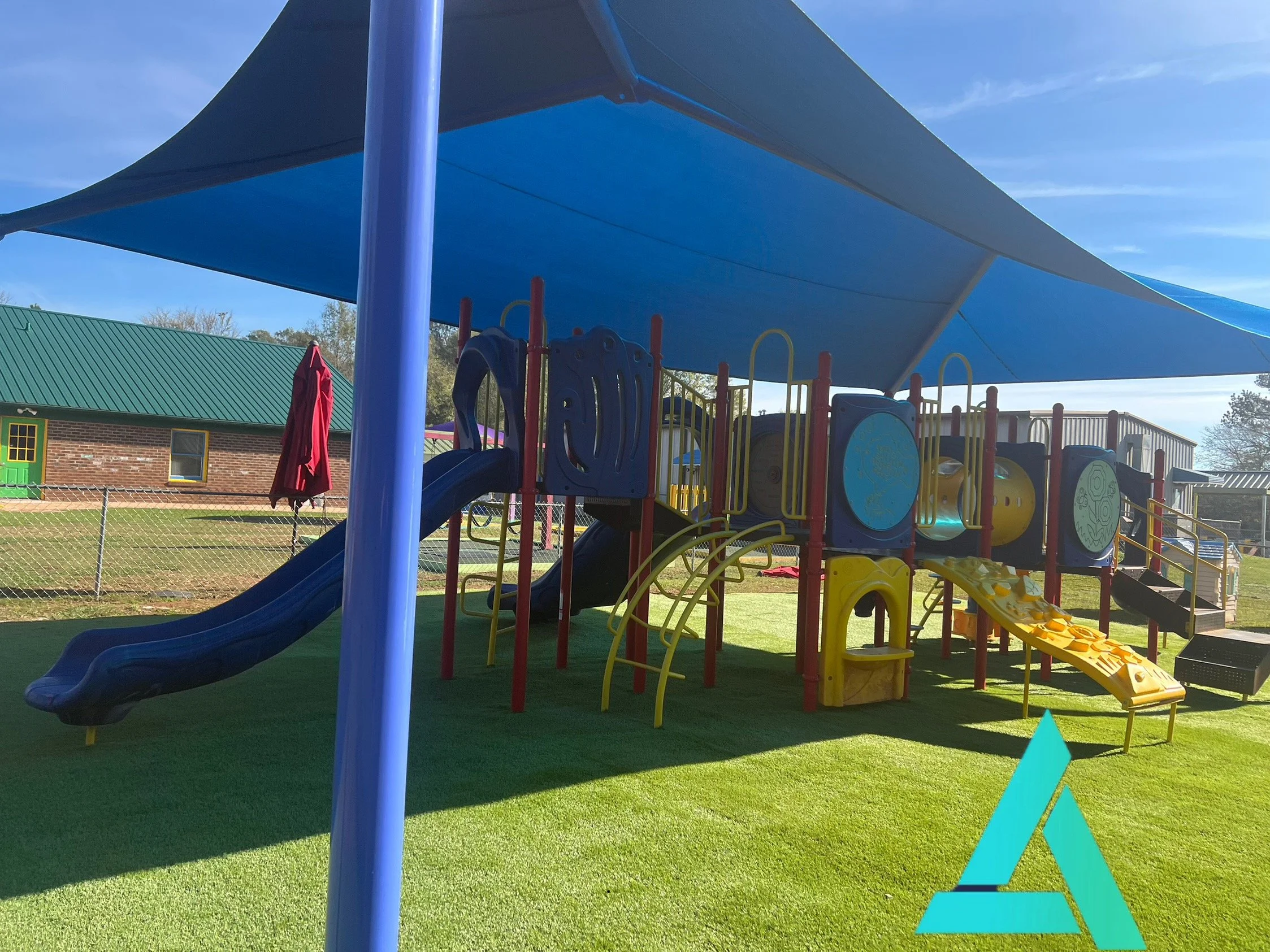 A colorful playground with slides, climbing structures, and a large blue shade canopy. There is a brick building with green roof and yellow window edges in the background, and the ground is covered with green artificial turf.