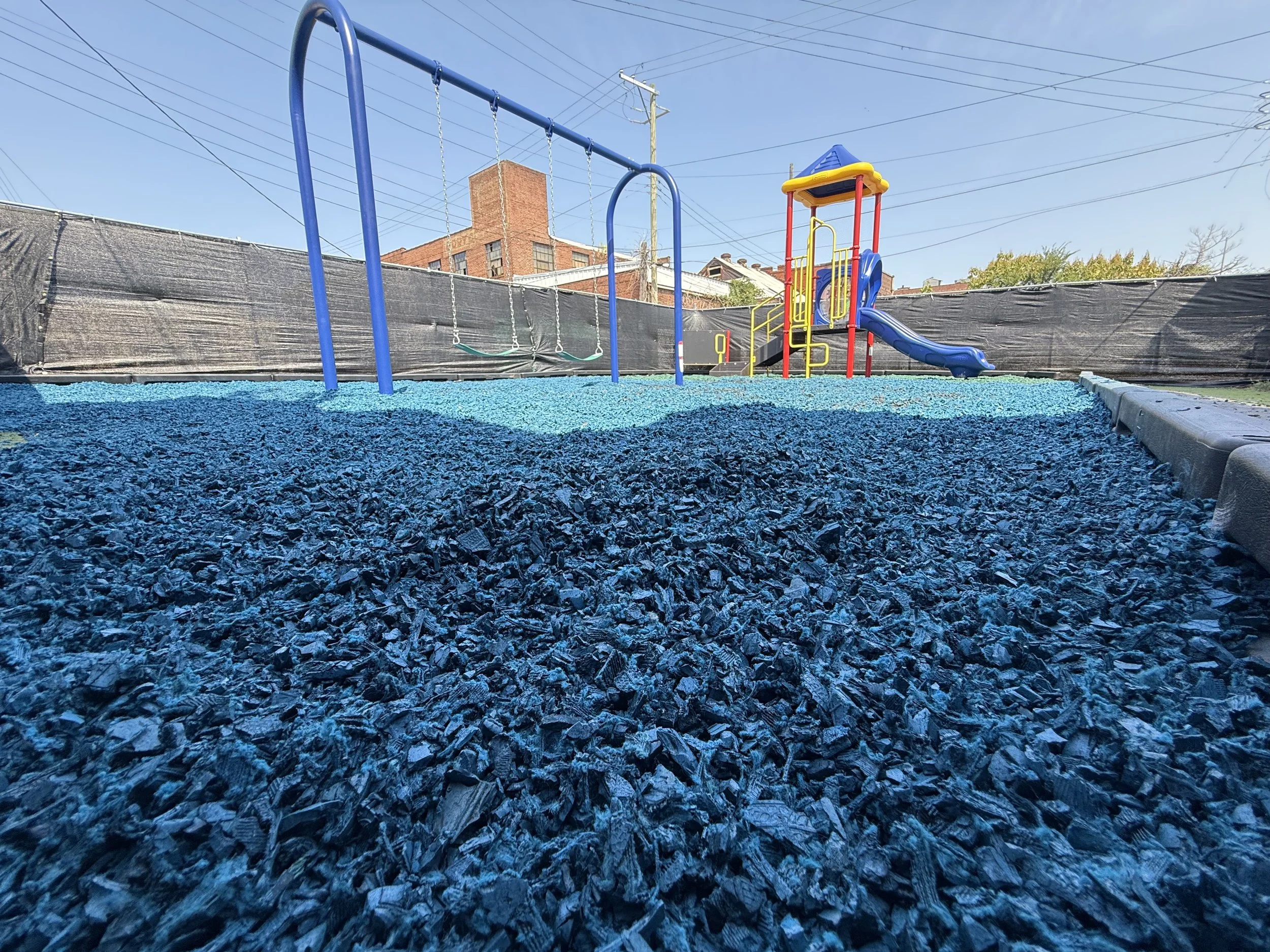Colorful playground with blue rubber ground, blue swing set, and slides, surrounded by a black fence and utility poles, under a clear sky.