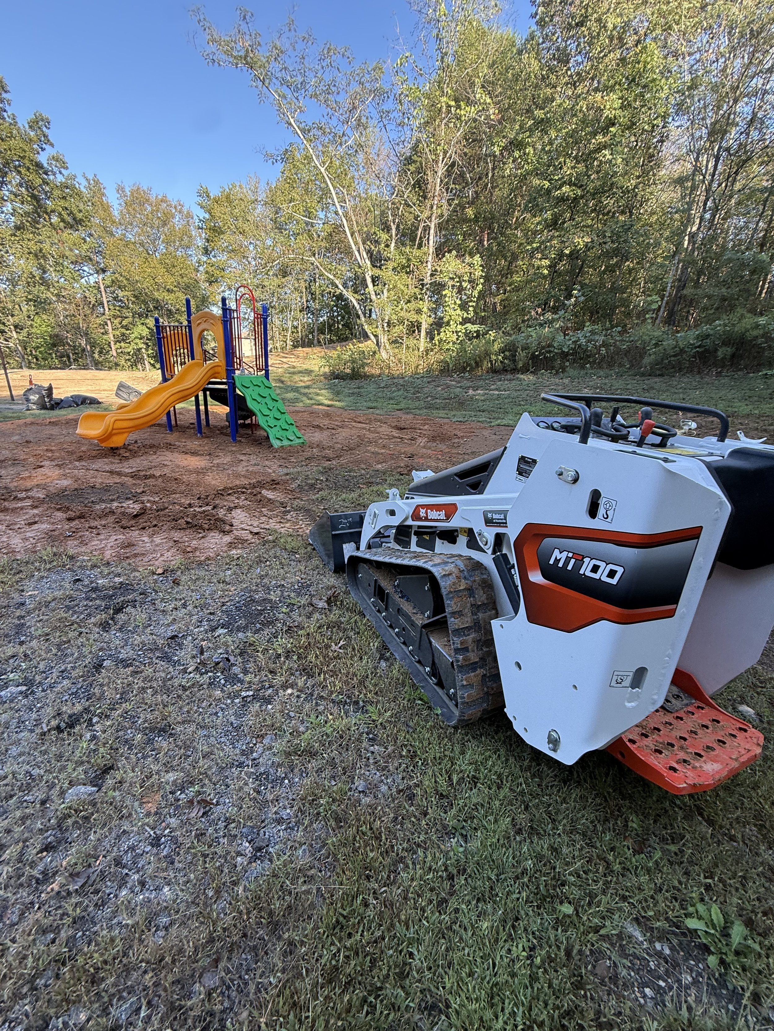 A small construction vehicle with tracks labeled 'MiT100' parked on grass and soil, with a playground structure featuring a yellow slide and climbing wall in the background, surrounded by trees and a clear blue sky.