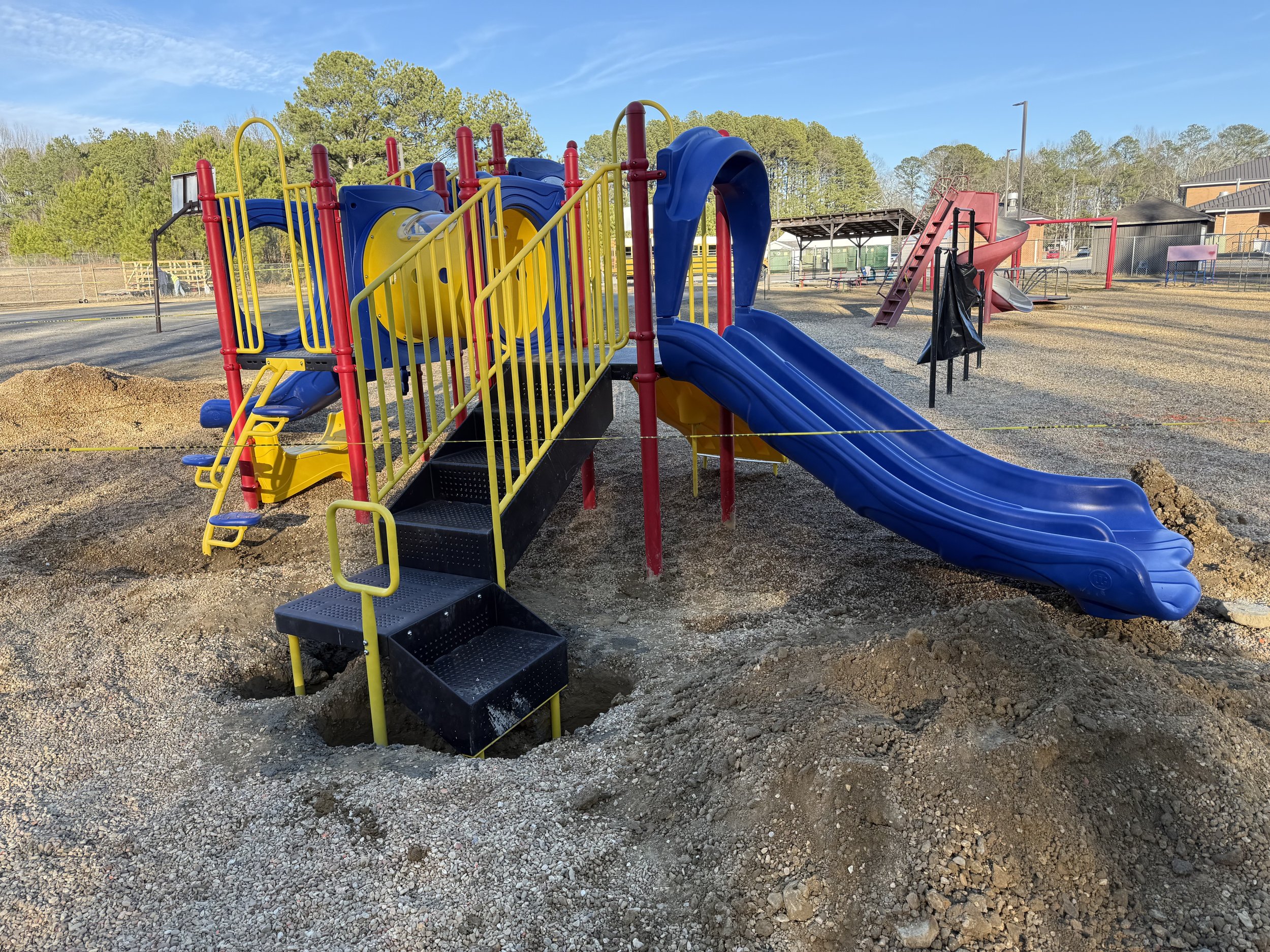 Colorful playground with blue slides, yellow railings, and red bars, under a clear blue sky, in a fenced outdoor area with trees in the background.