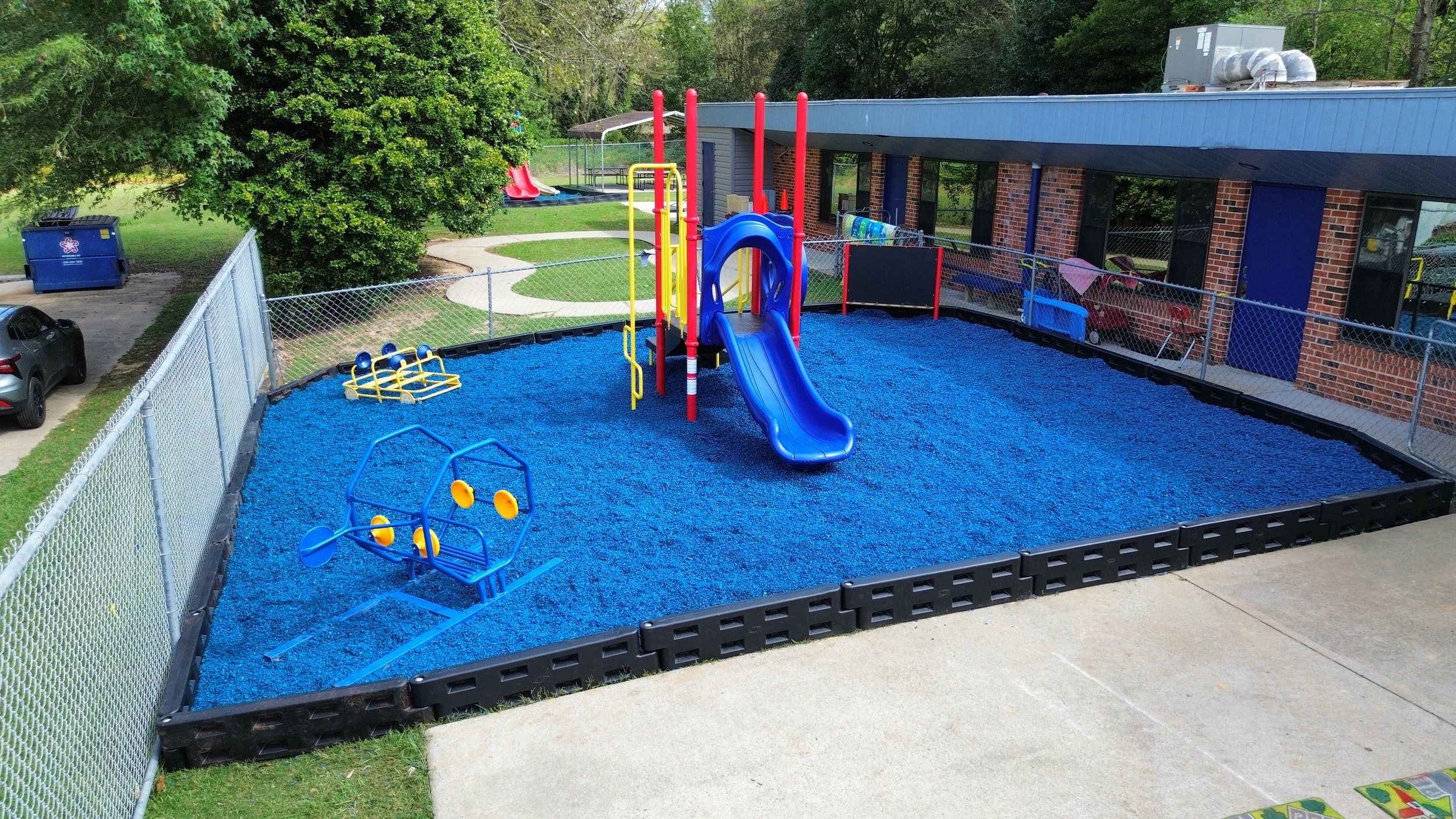 A fenced playground area with blue rubber playground mulch , a blue slide, yellow climbing structures, and small blue and yellow spinning seats. In the background, there are trees, a building with brick walls, patio furniture, and a playset with slid